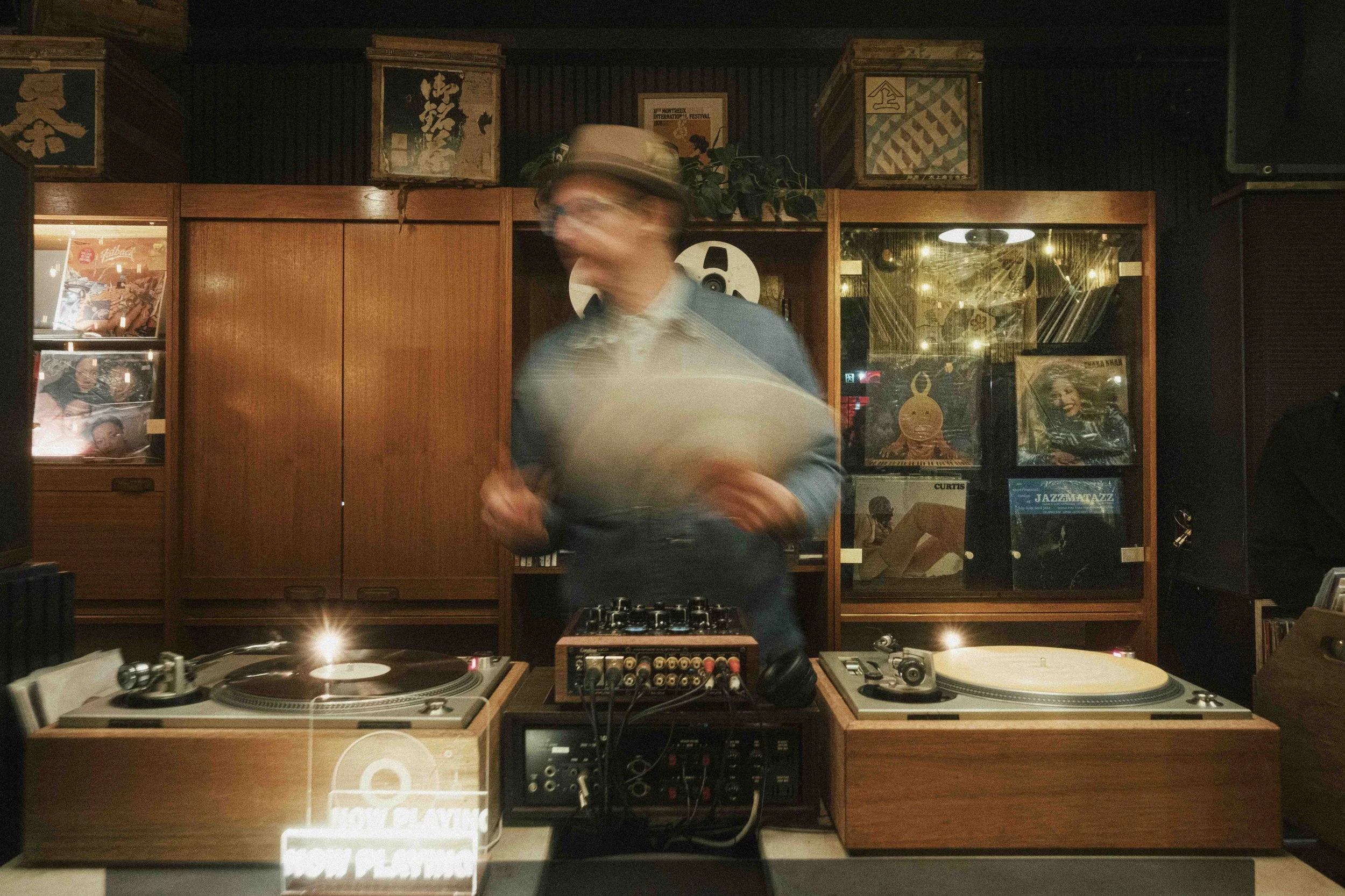 A DJ is performing behind turntables in a dimly lit room with vinyl records on display in the background.