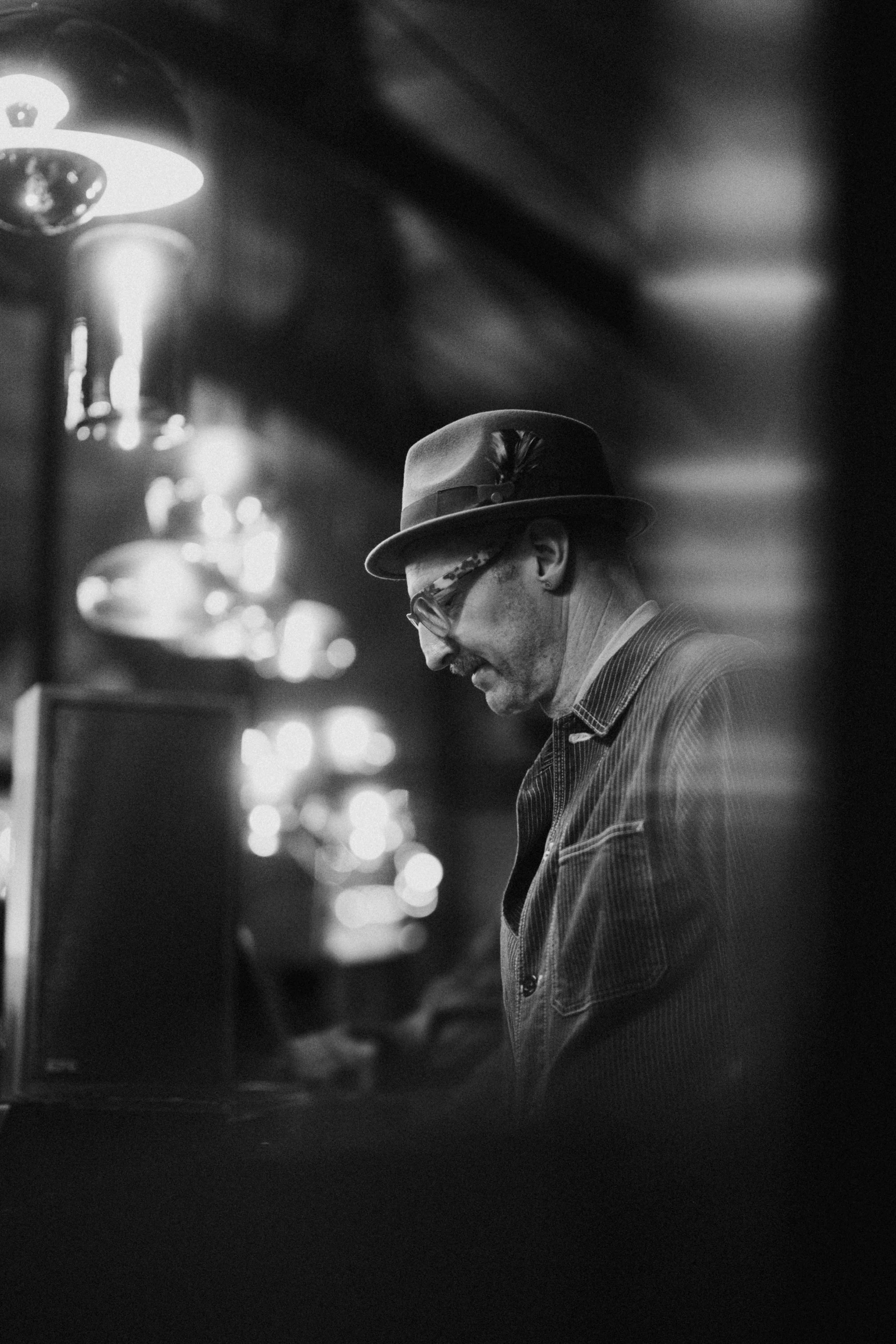 A black-and-white photo of a man wearing a fedora hat, sunglasses, and a striped jacket, looking down while playing a piano or keyboard in a dimly lit room with decorative lights.