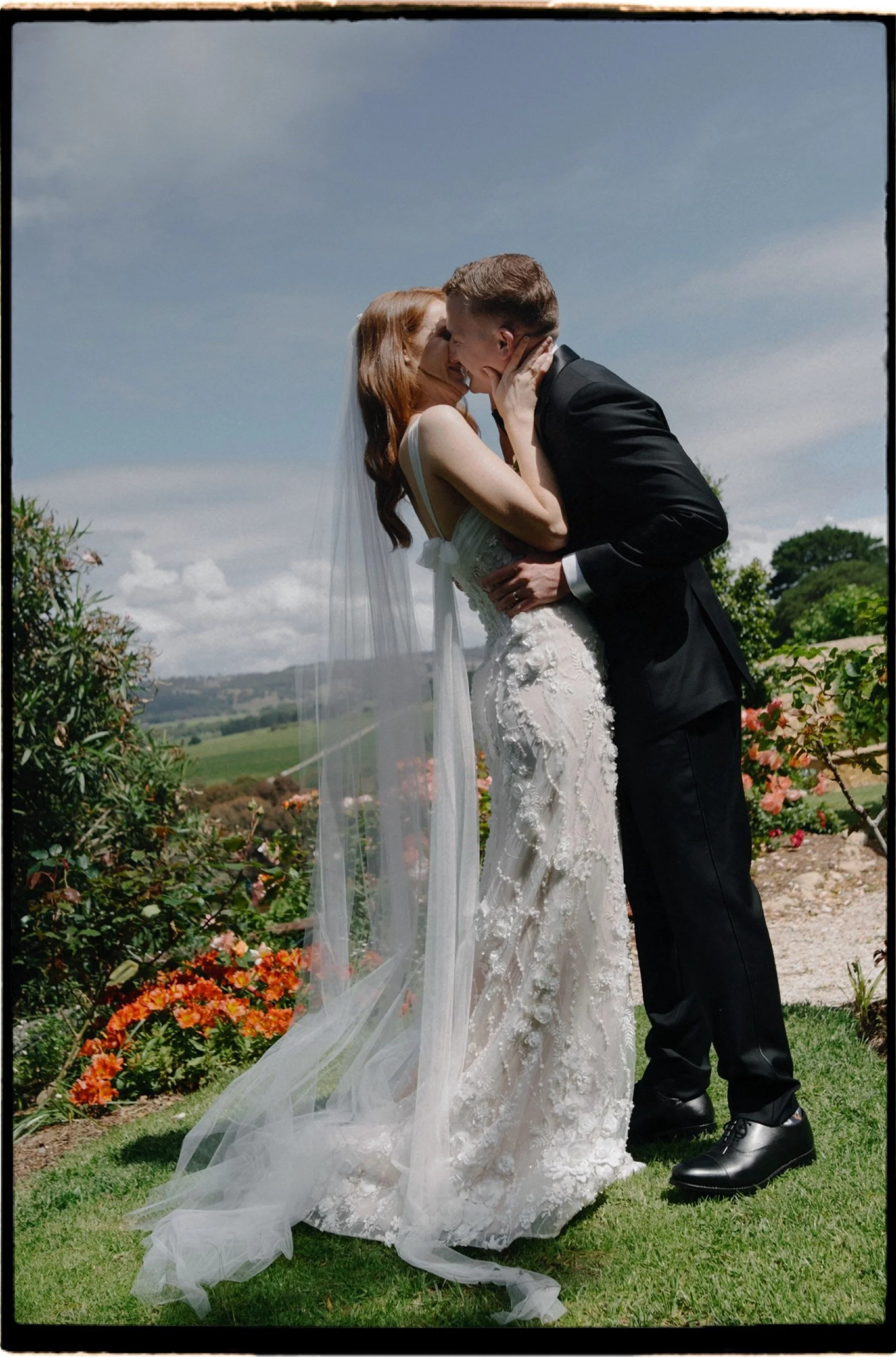 A bride and groom sharing a kiss outside in a garden with flowers and green hills in the background.
