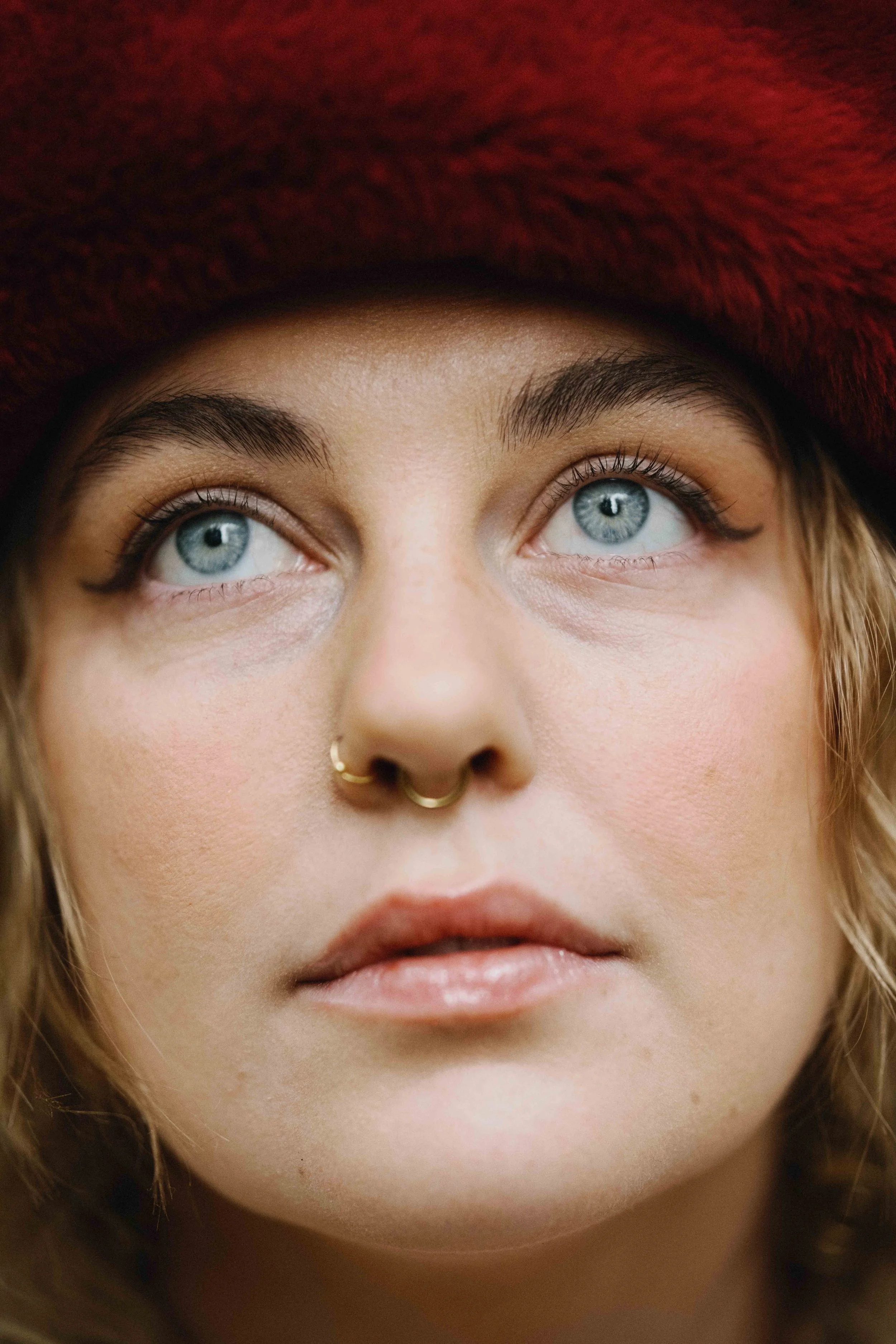 Close-up of a woman's face looking upwards. She has blue eyes, a septum piercing, and is wearing a red, fuzzy hat.
