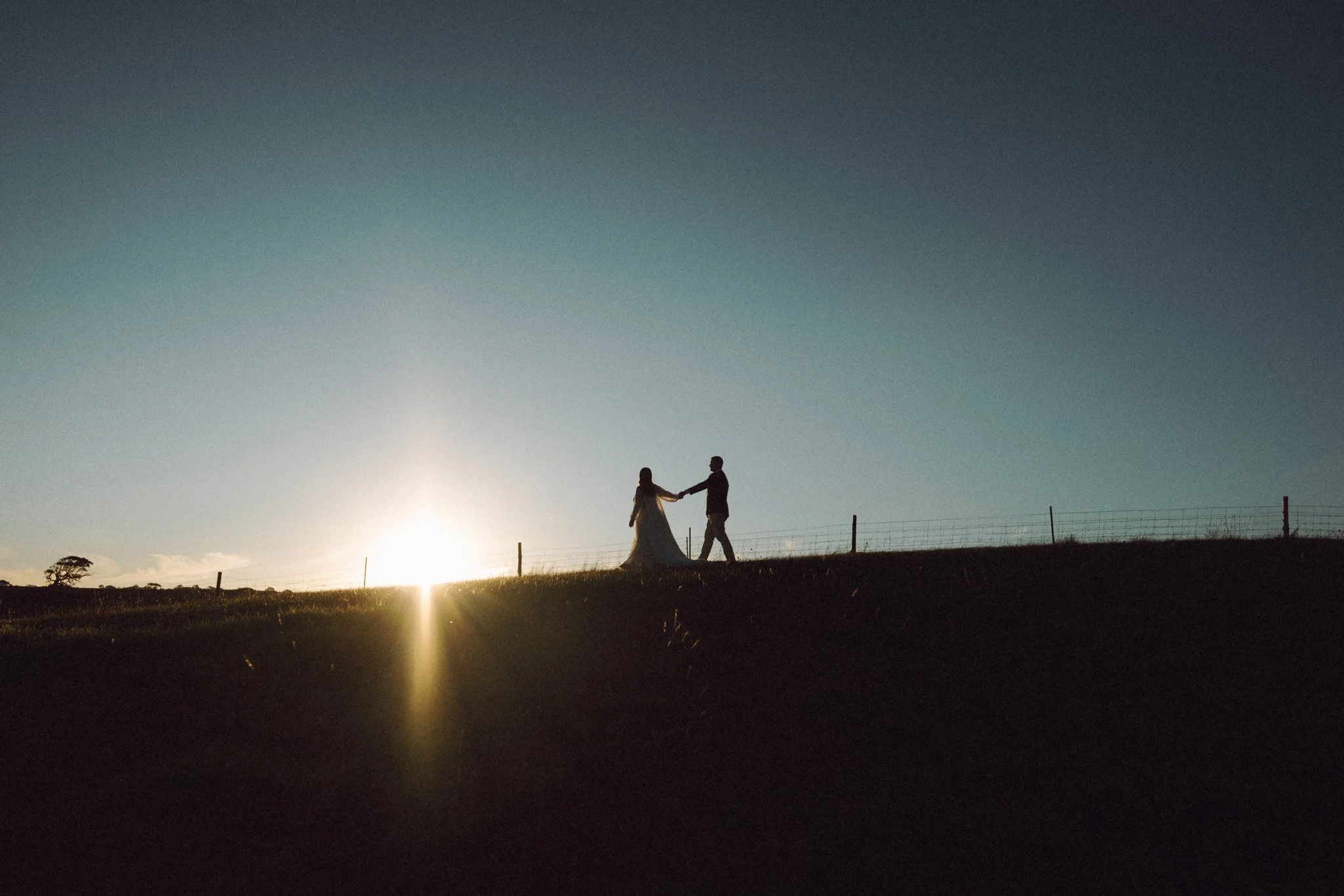 A silhouette of a couple holding hands and dancing outdoors during sunset, with a fence and open landscape in the background.