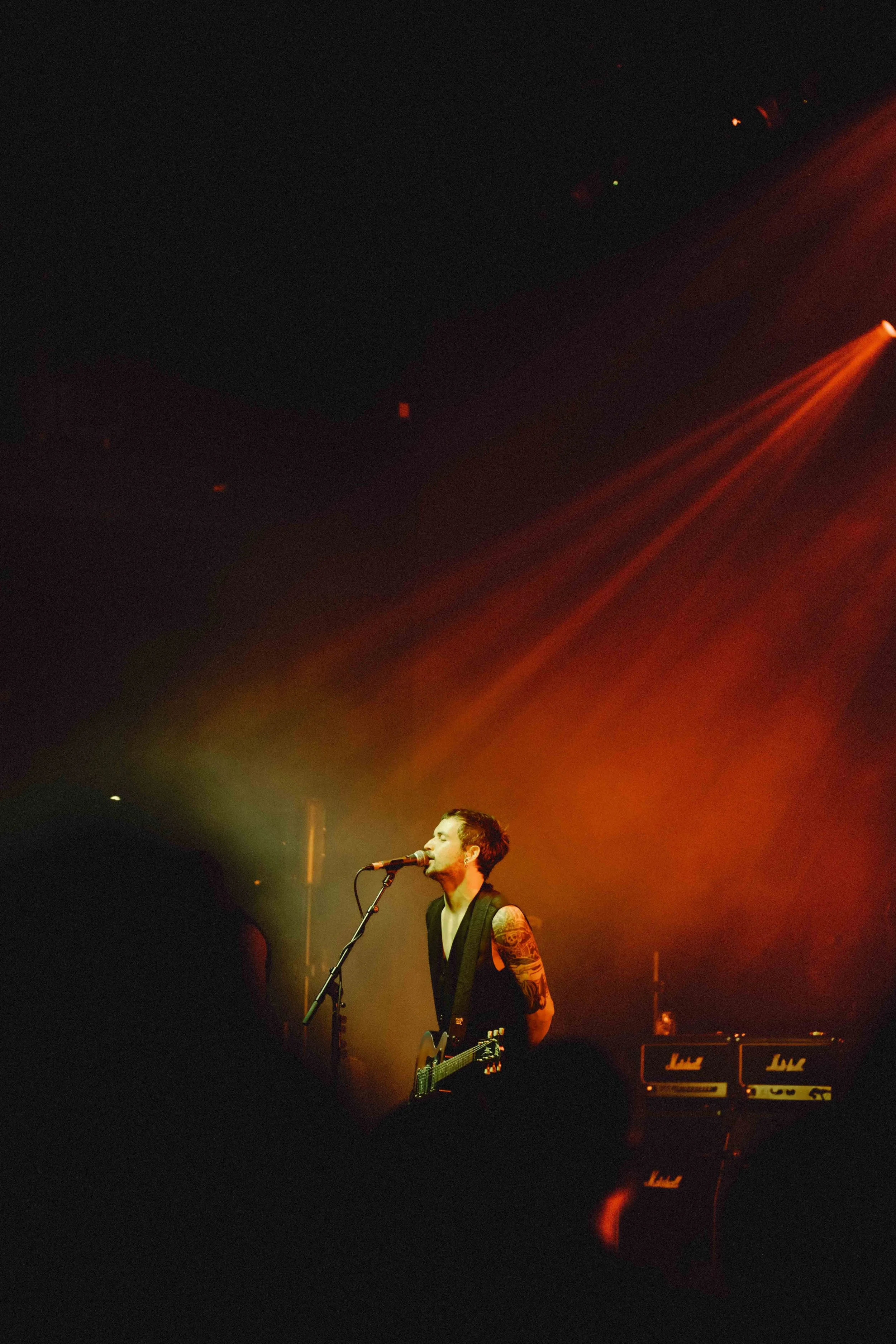 A male singer with tattoos on his arm performing on stage with a microphone and guitar in front of an audience, with orange stage lighting.