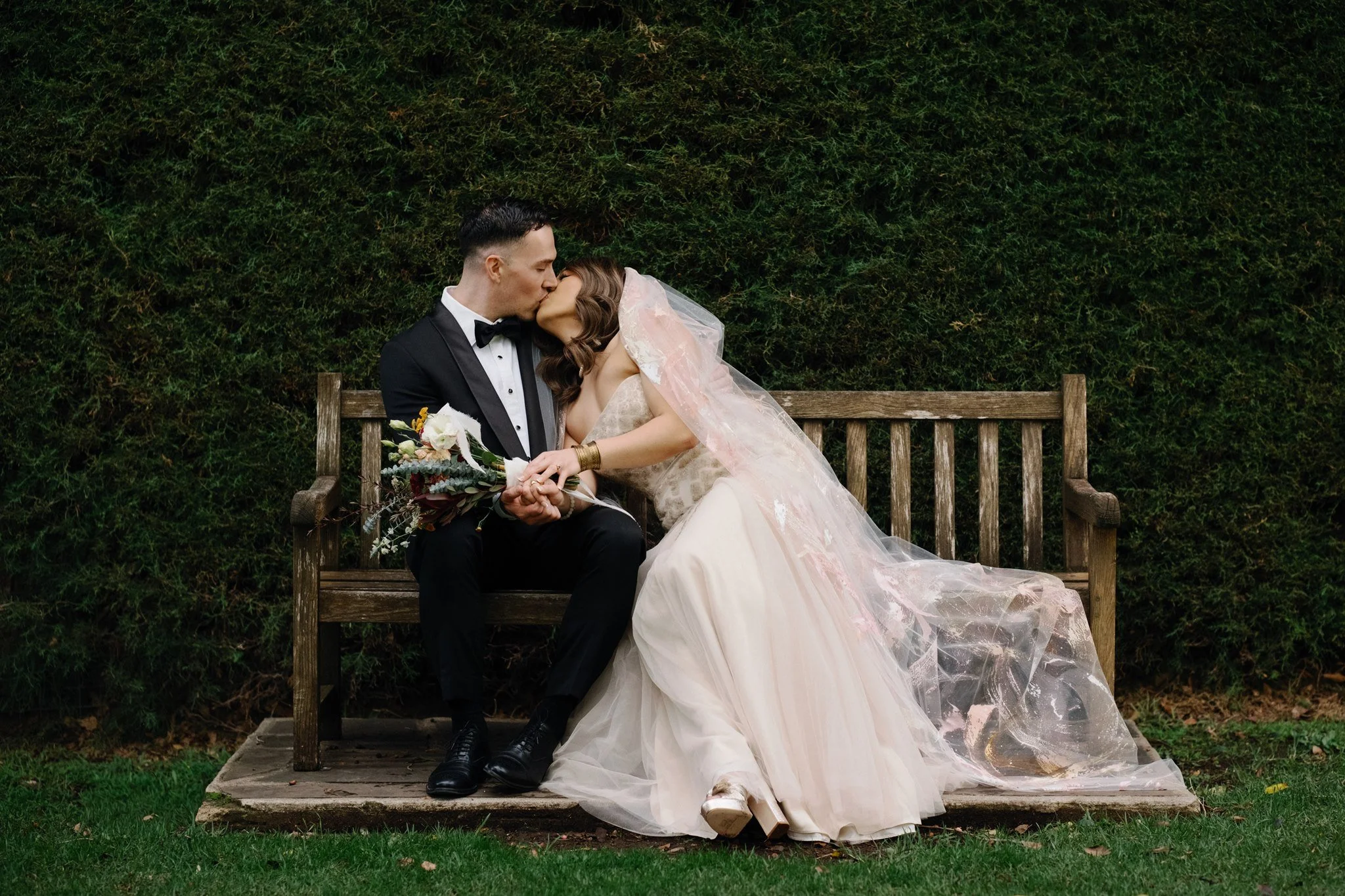 A bride and groom sharing a kiss on a park bench. The bride is wearing a wedding gown with a veil and holding a bouquet, while the groom is in a tuxedo. They are sitting outdoors against a leafy green backdrop.