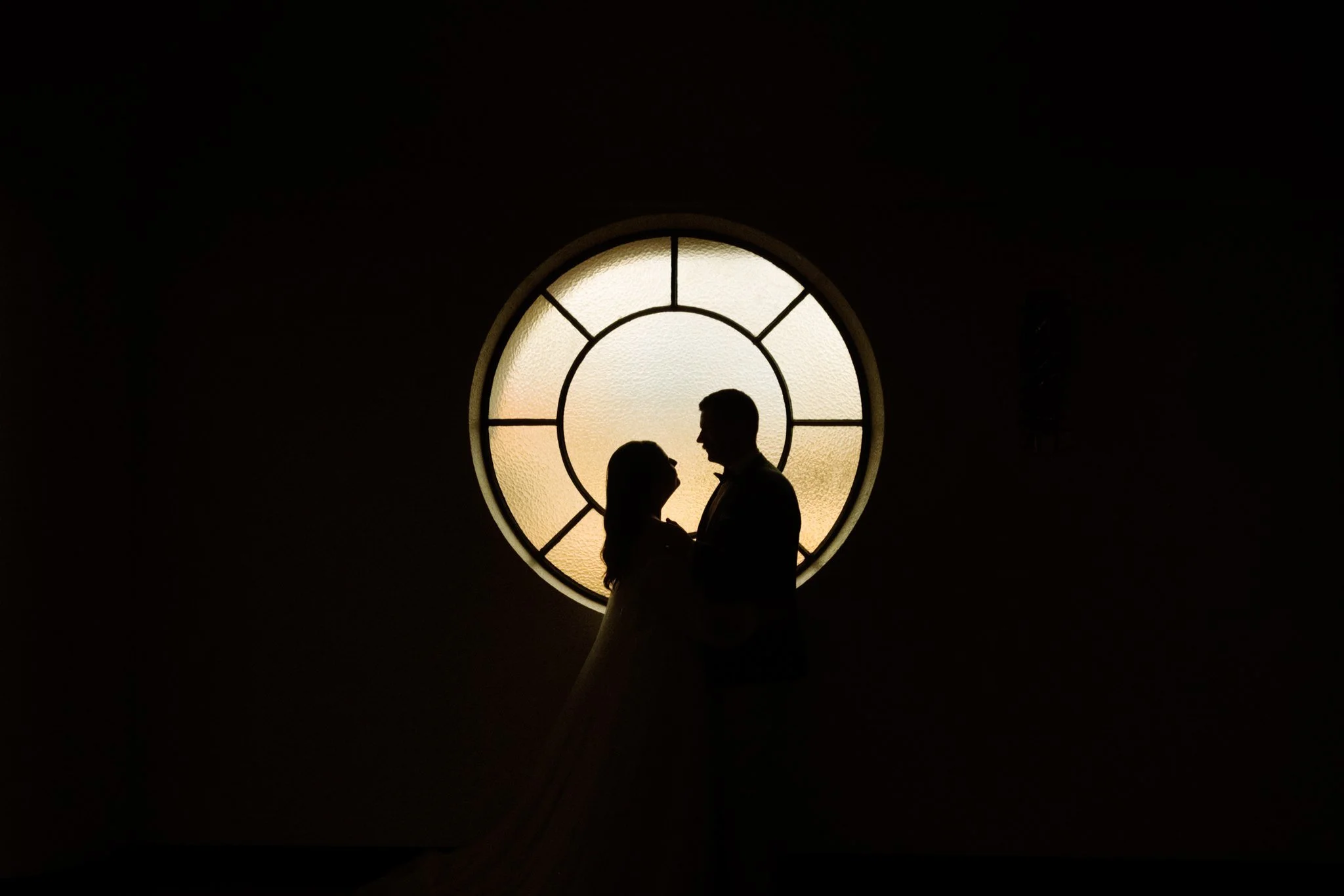Silhouette of a couple standing close together in front of a round window with stained glass, backlit by light coming through the window.