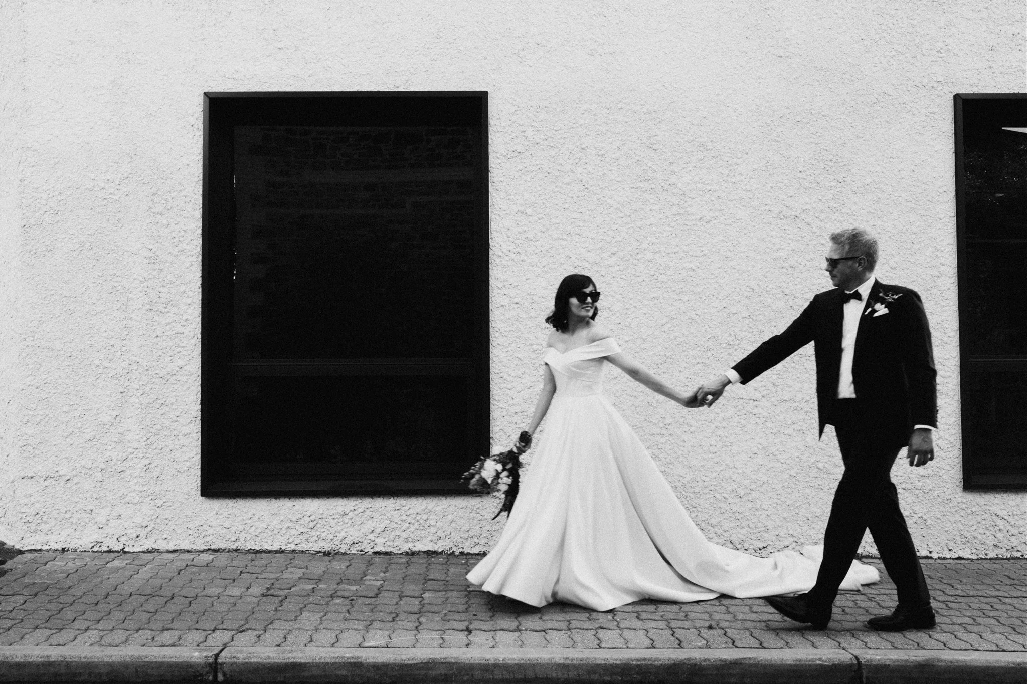 A black-and-white photo of a bride in a wedding dress holding a bouquet and a groom in a tuxedo holding hands, walking on a sidewalk in front of a textured wall with dark windows.