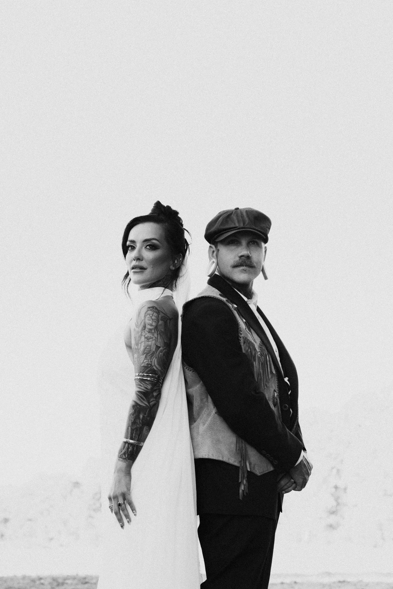 A black and white photo of a woman and man standing back to back outdoors, with an open sky in the background. The woman has tattoos on her arms and shoulder, and the man is wearing a flat cap and has a mustache.