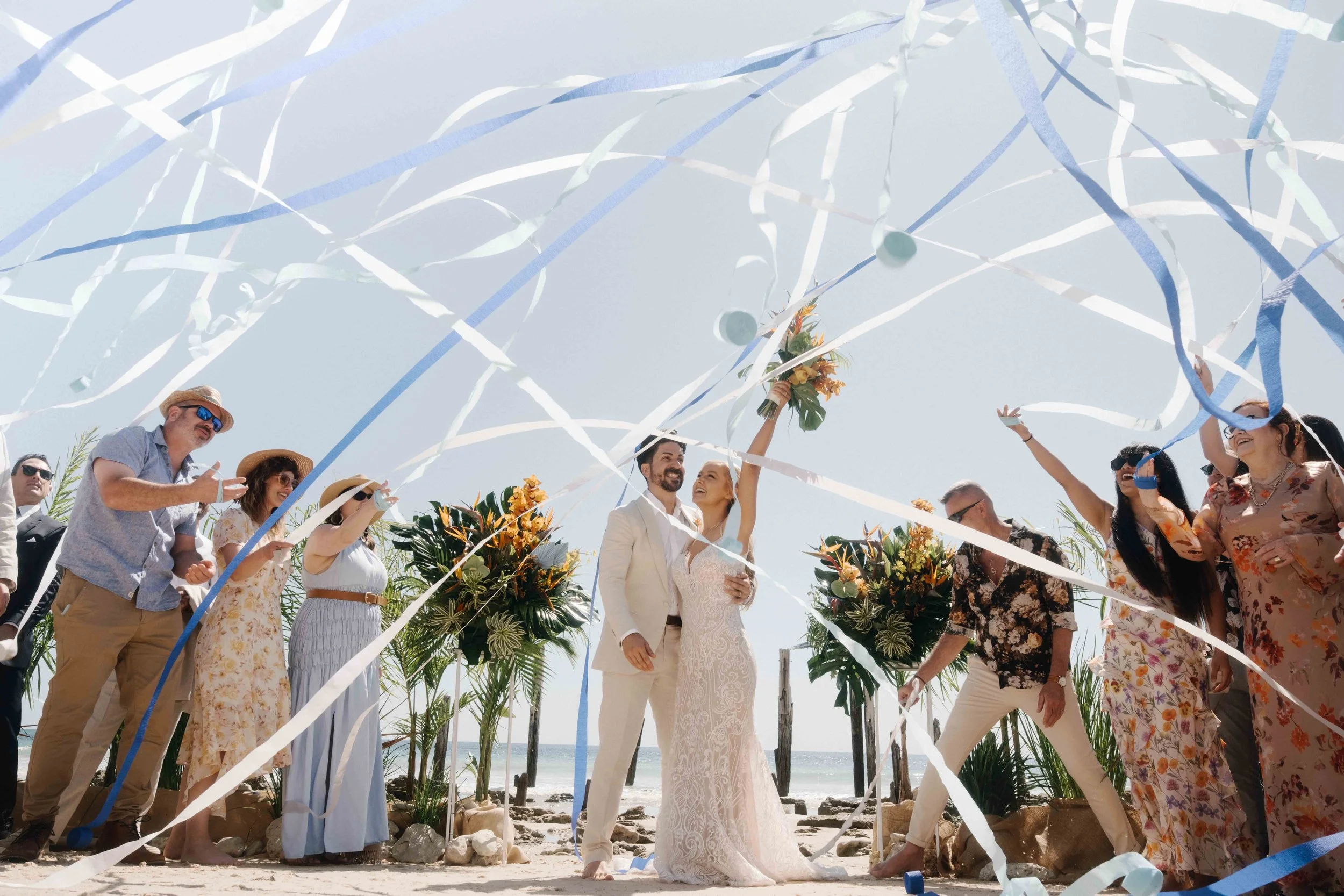 A beach wedding ceremony with a bride and groom surrounded by friends and family, celebrating with streamers and flowers.