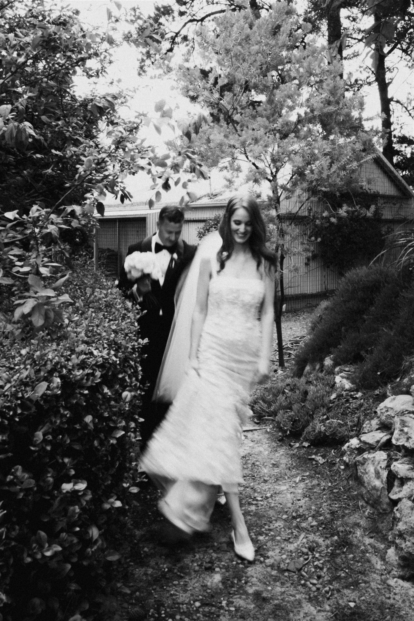 A black and white photo of a bride in a wedding gown and a groom carrying a bouquet of flowers walking on a garden path surrounded by trees and bushes.