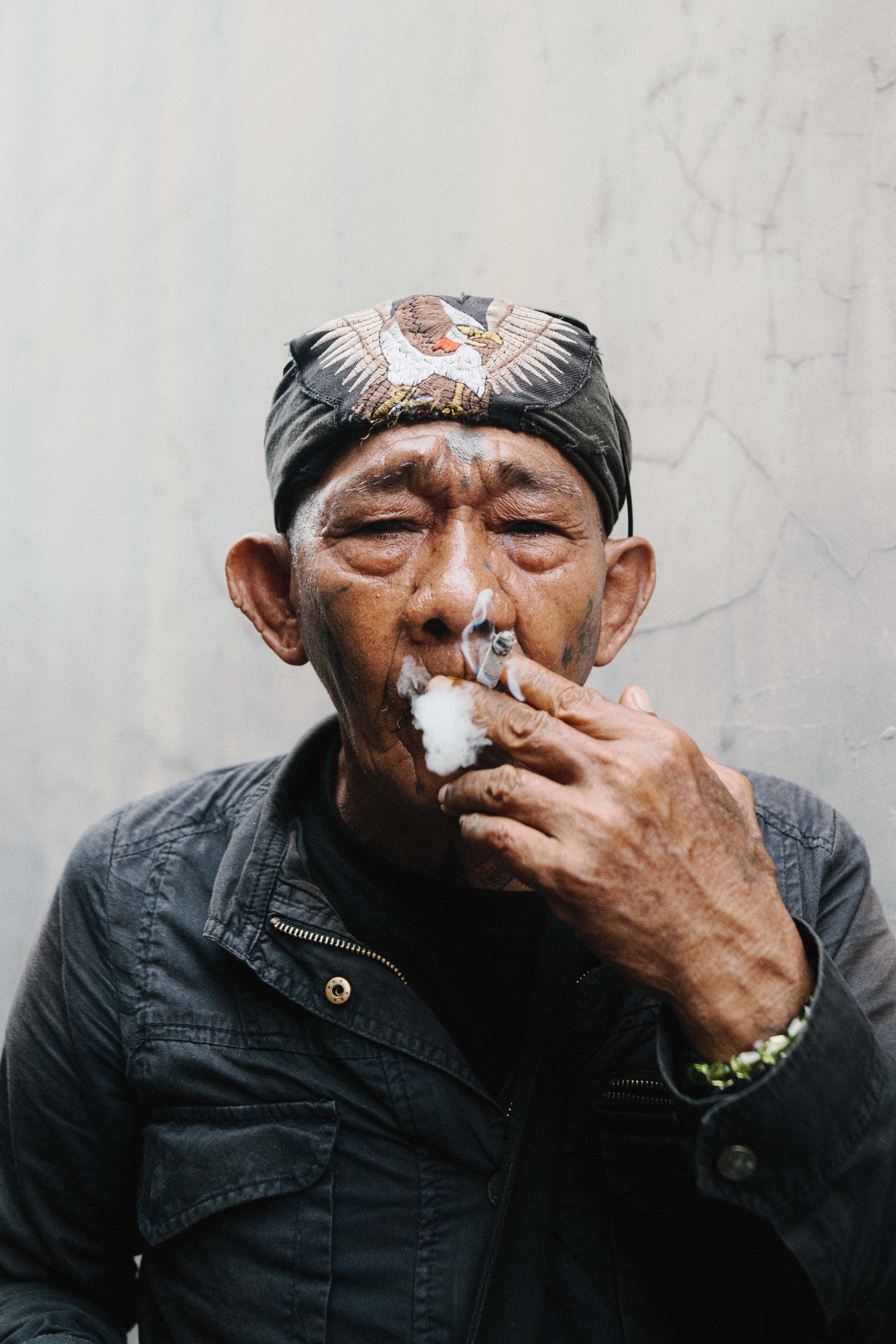 A man with tattoos on his face and hand, wearing a black headband with an eagle embroidery, smokes a cigarette, holding it with his left hand. The background is plain and light-colored.