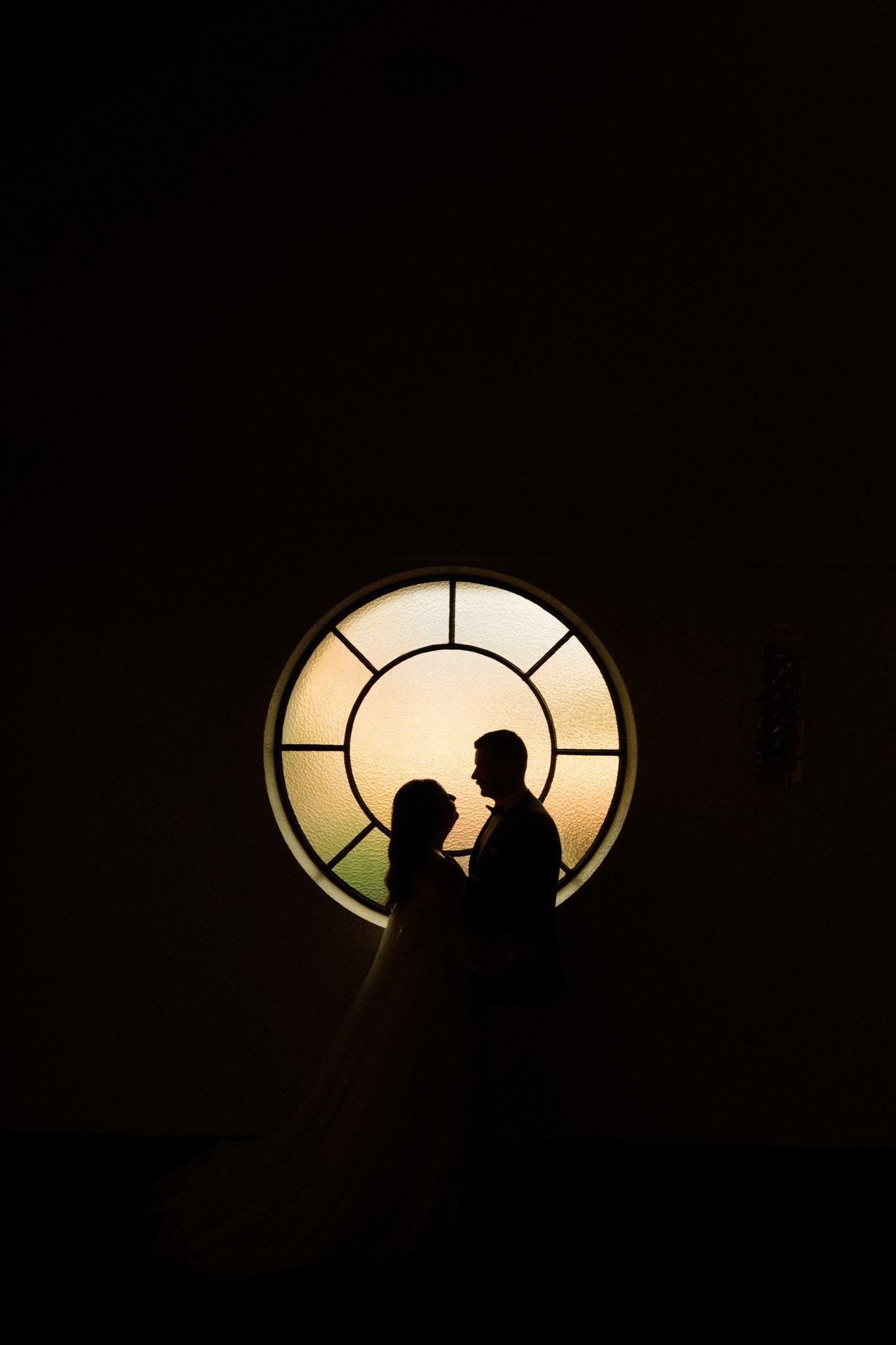 Silhouette of a couple facing each other in front of a large circular window with stained glass design, during sunset or sunrise.