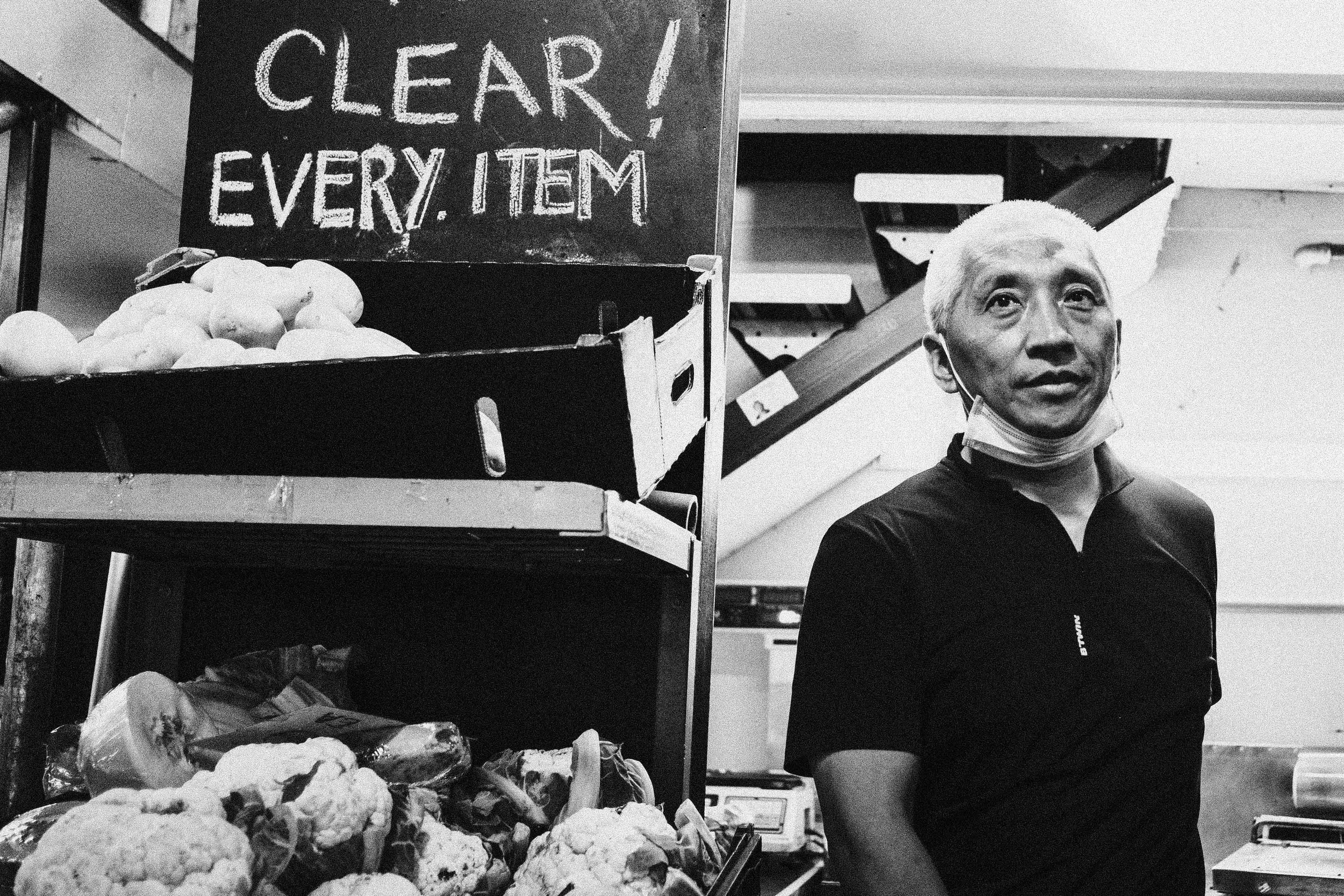 A man with platinum blonde hair and a face mask around his neck is standing next to a produce display in a grocery store. The display has a sign that reads "CLEAR! EVERY ITEM" and holds various vegetables, including cauliflower and what appear to be 