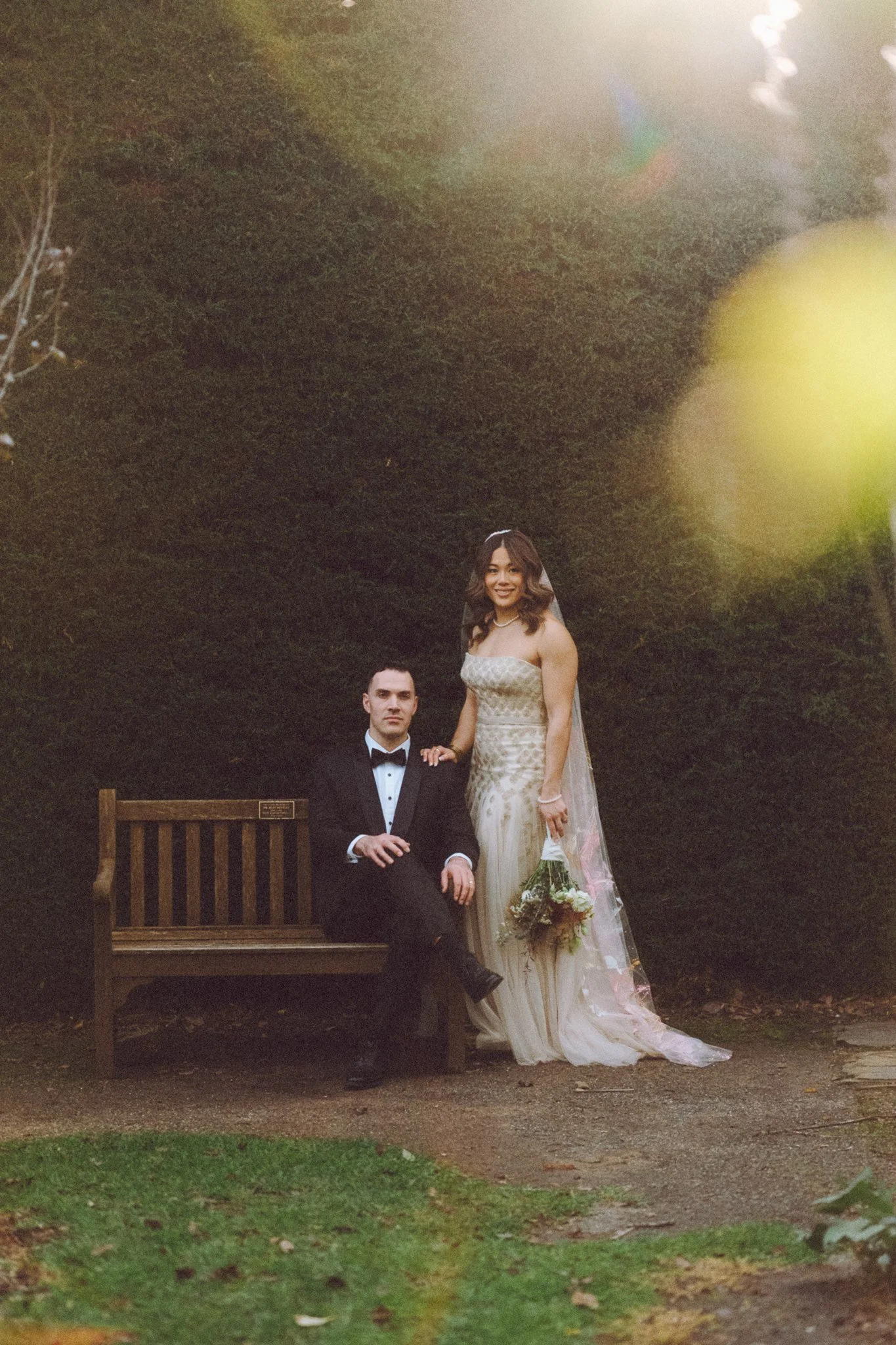 A couple dressed in wedding attire posing outdoors at night. The woman is standing, wearing a white wedding dress and holding a bouquet. The man is sitting on a wooden bench, wearing a black tuxedo with a bow tie. There are trees and greenery in the 