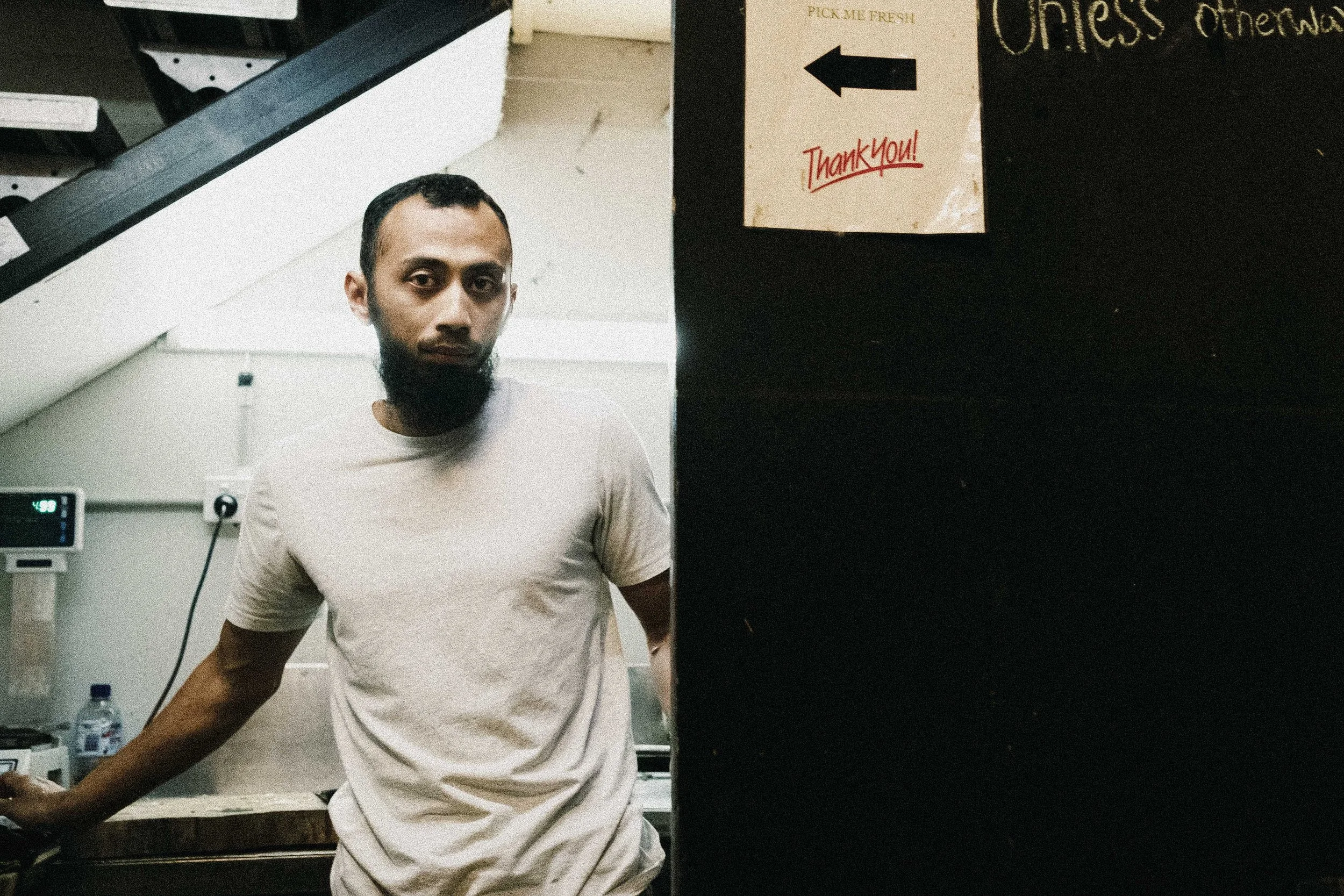 A man with a beard wearing a white t-shirt standing in a room with white and black walls, next to a black surface with a sign pointing left that says "Pick Me Fresh" and "Thank You!"