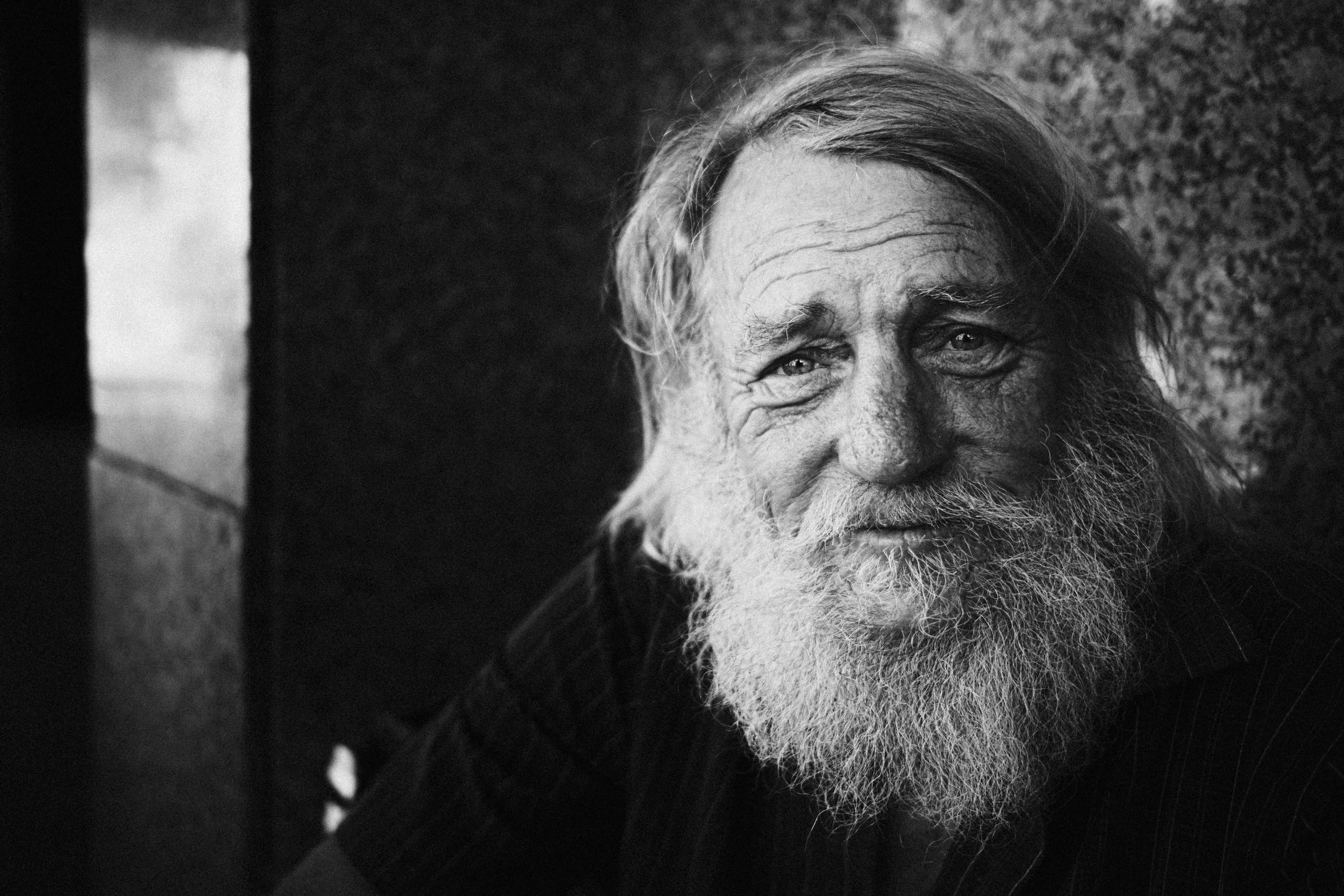 A black and white photo of an elderly man with a long, white beard and long, unkempt hair, looking directly at the camera with a gentle expression.