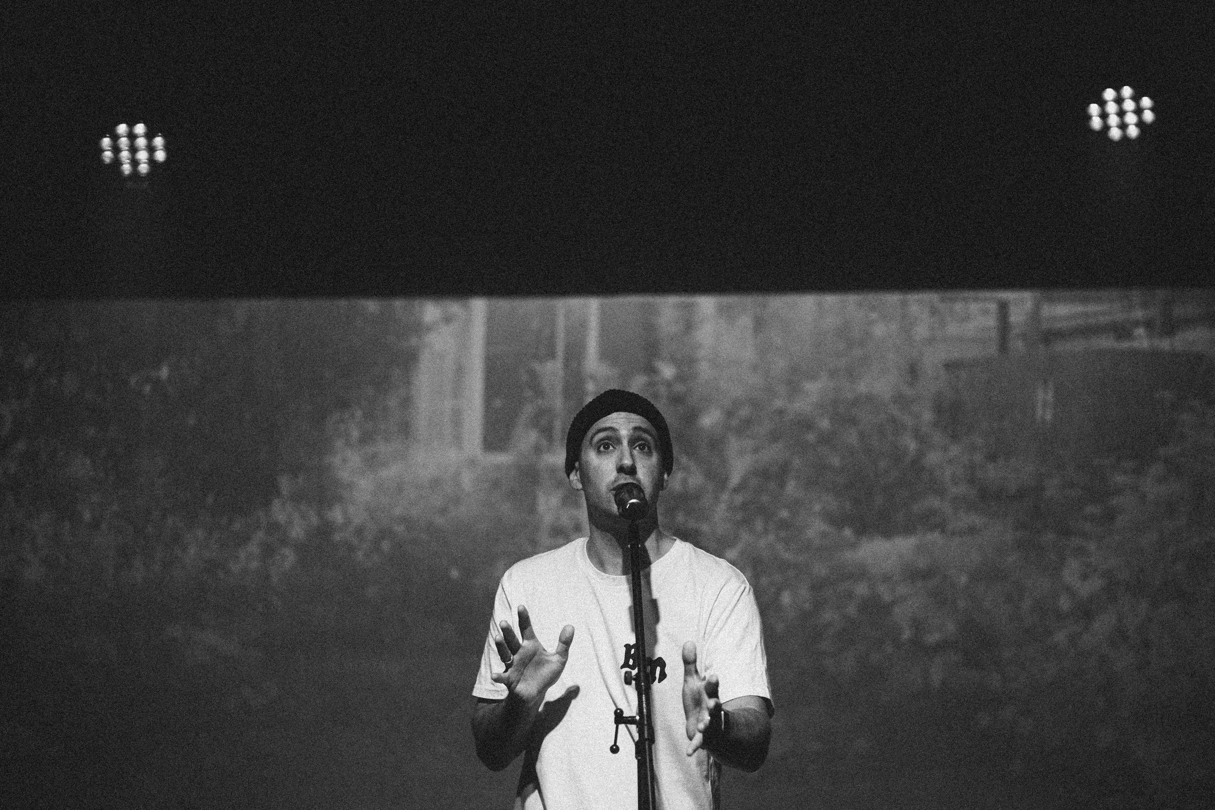 A person with short hair, wearing a beanie and a white t-shirt, speaking into a microphone on a stand with a serious expression, in front of a forest background, black and white photo.