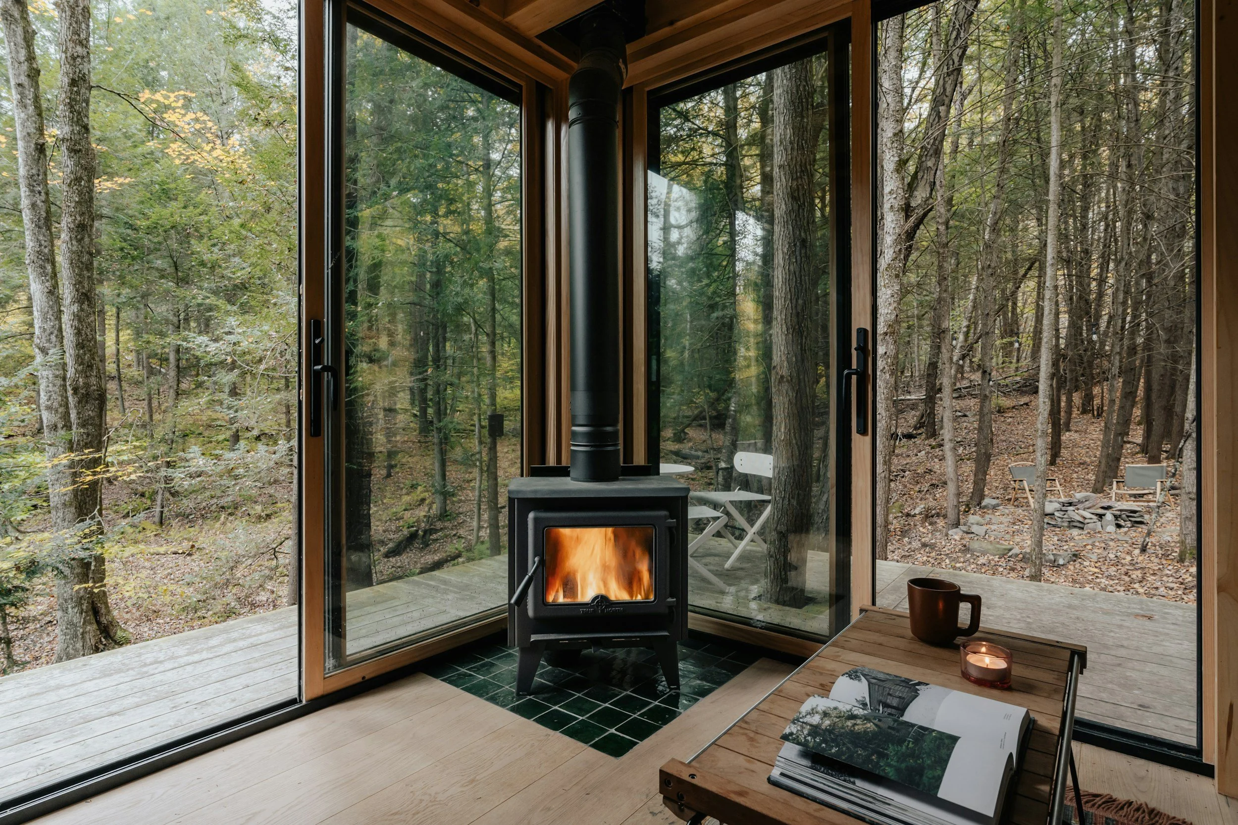 A cozy room with floor-to-ceiling glass doors opening to a forest outdoor space. There is a wood-burning stove with a fire inside, situated on a green tiled base, near a wooden table that holds an open magazine, a candle, and a ceramic mug. Outside, visible through the glass, is a wooden deck with outdoor chairs, and a wooded area with trees.