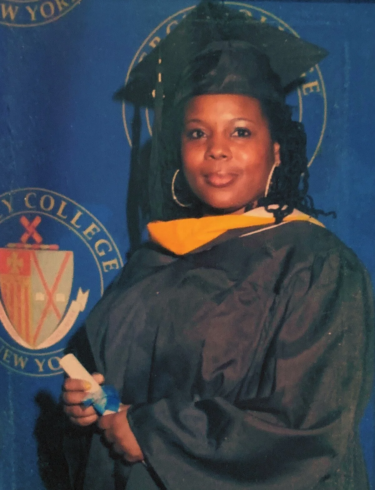 A woman in a graduation cap and gown standing in front of a blue backdrop with the emblems of New York City College.