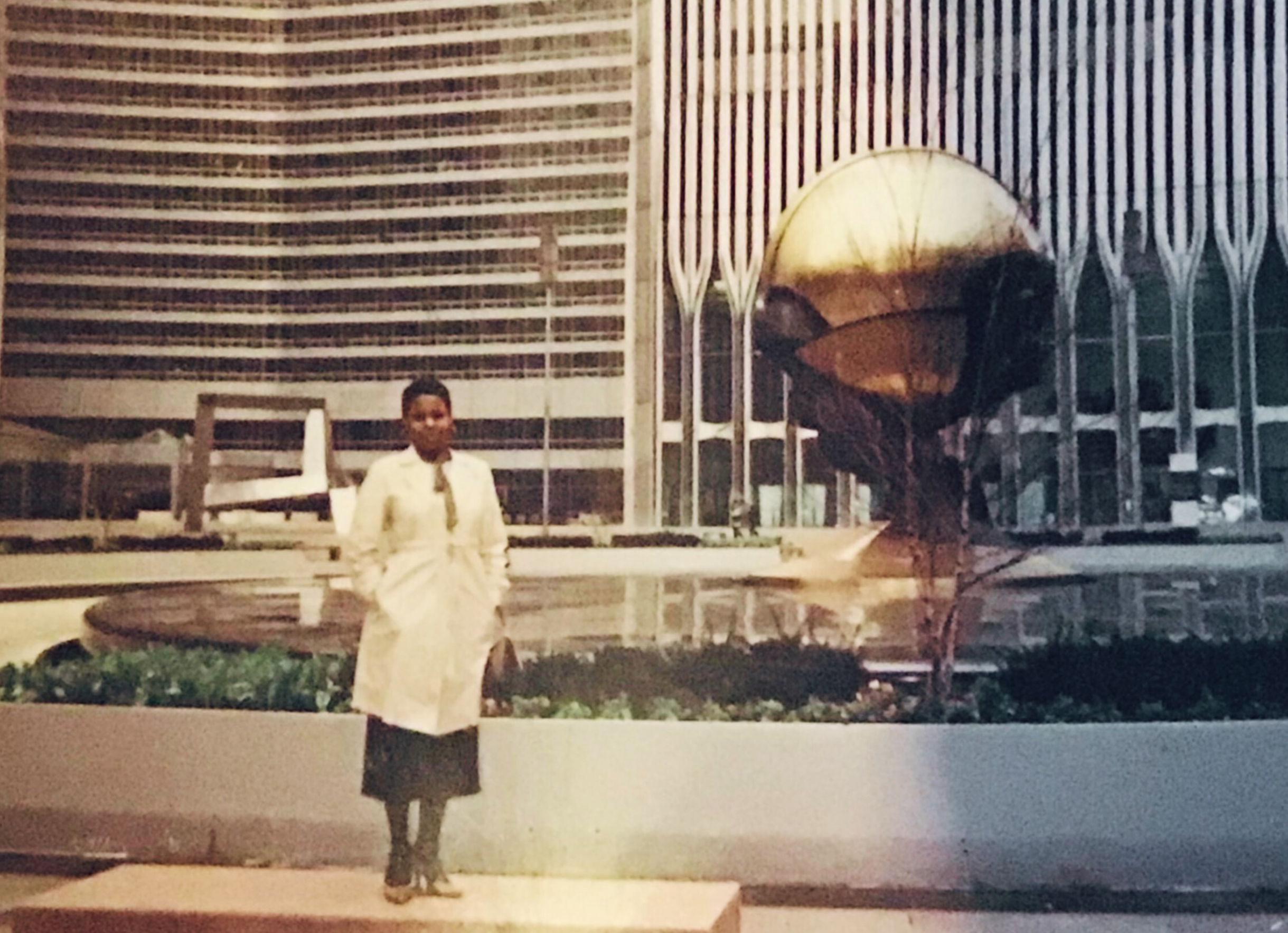 A woman in a white coat and black skirt standing in front of a modern building with large glass windows and a metallic sculpture resembling a ship's bow.