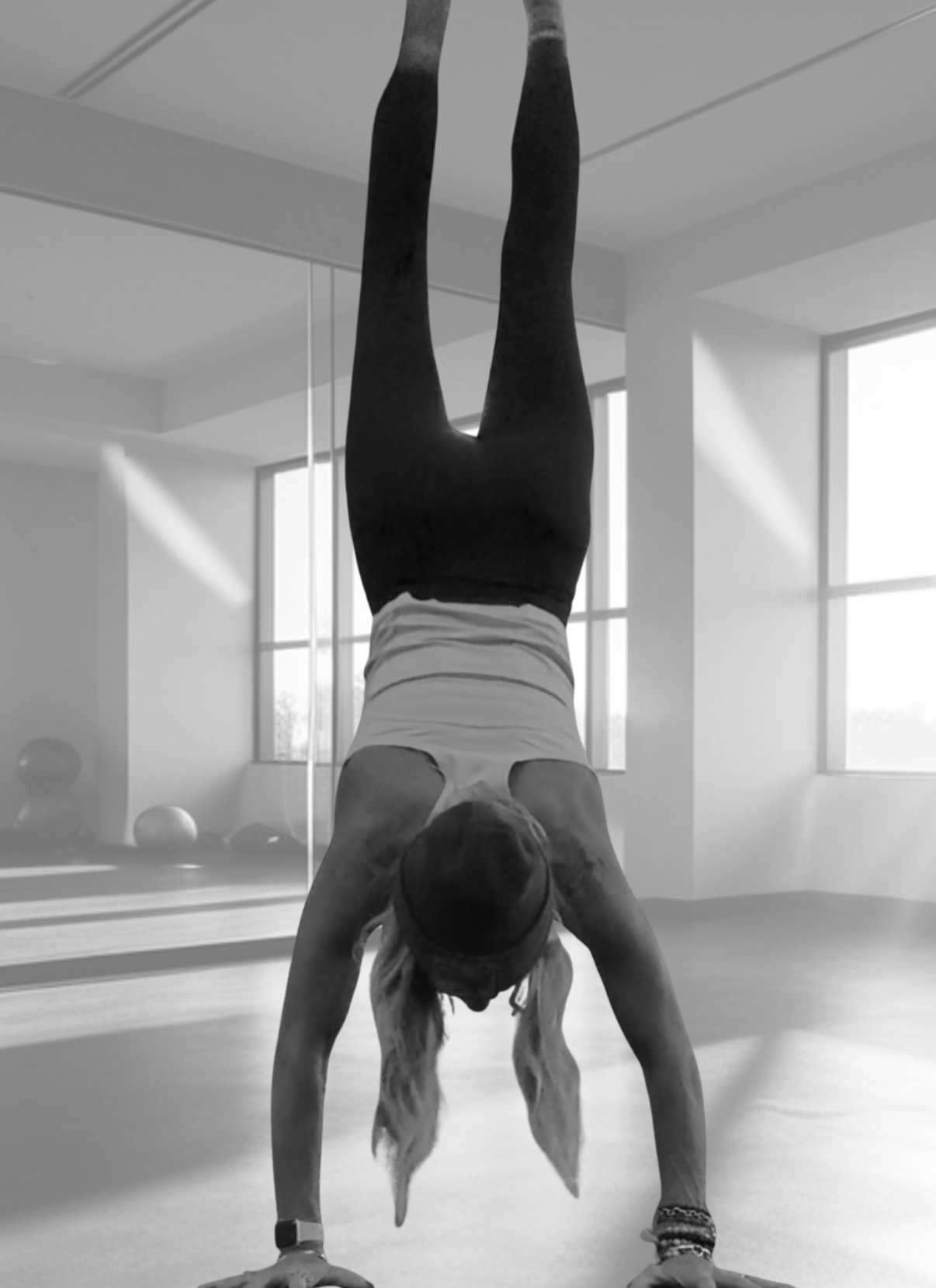 A woman performing a handstand in a brightly lit room with large windows and exercise balls in the background.