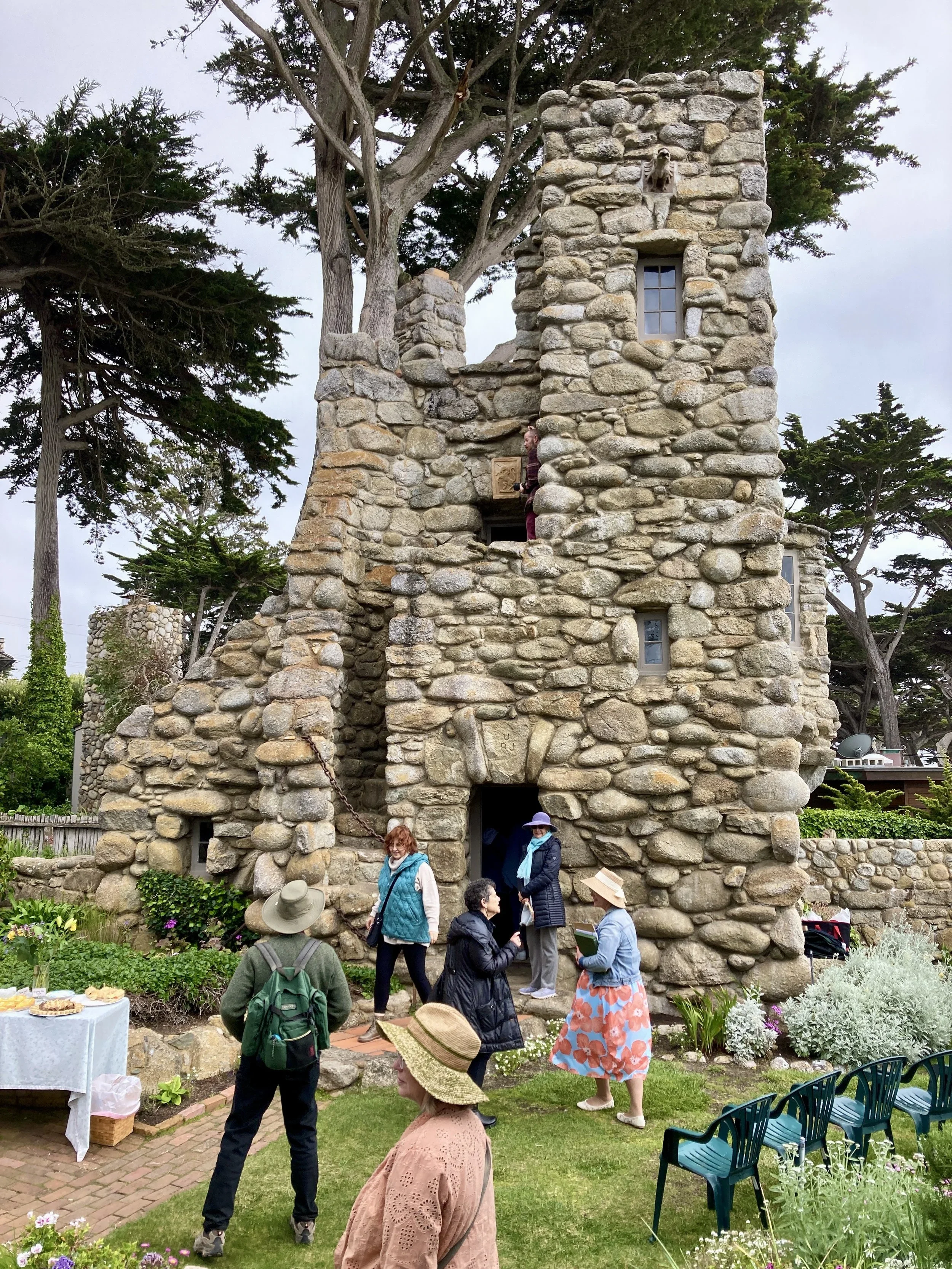 A group of people gathered around a stone tower with a rustic appearance, featuring small windows and a small opening at the bottom. Some people are on the ground, while one person stands on a ledge higher up in the tower. The scene is set in a garde