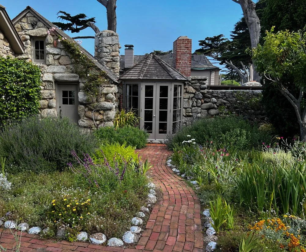 A stone cottage with a brick chimney and a small porch, surrounded by lush garden with a brick pathway, flowering plants, and trees under a blue sky.