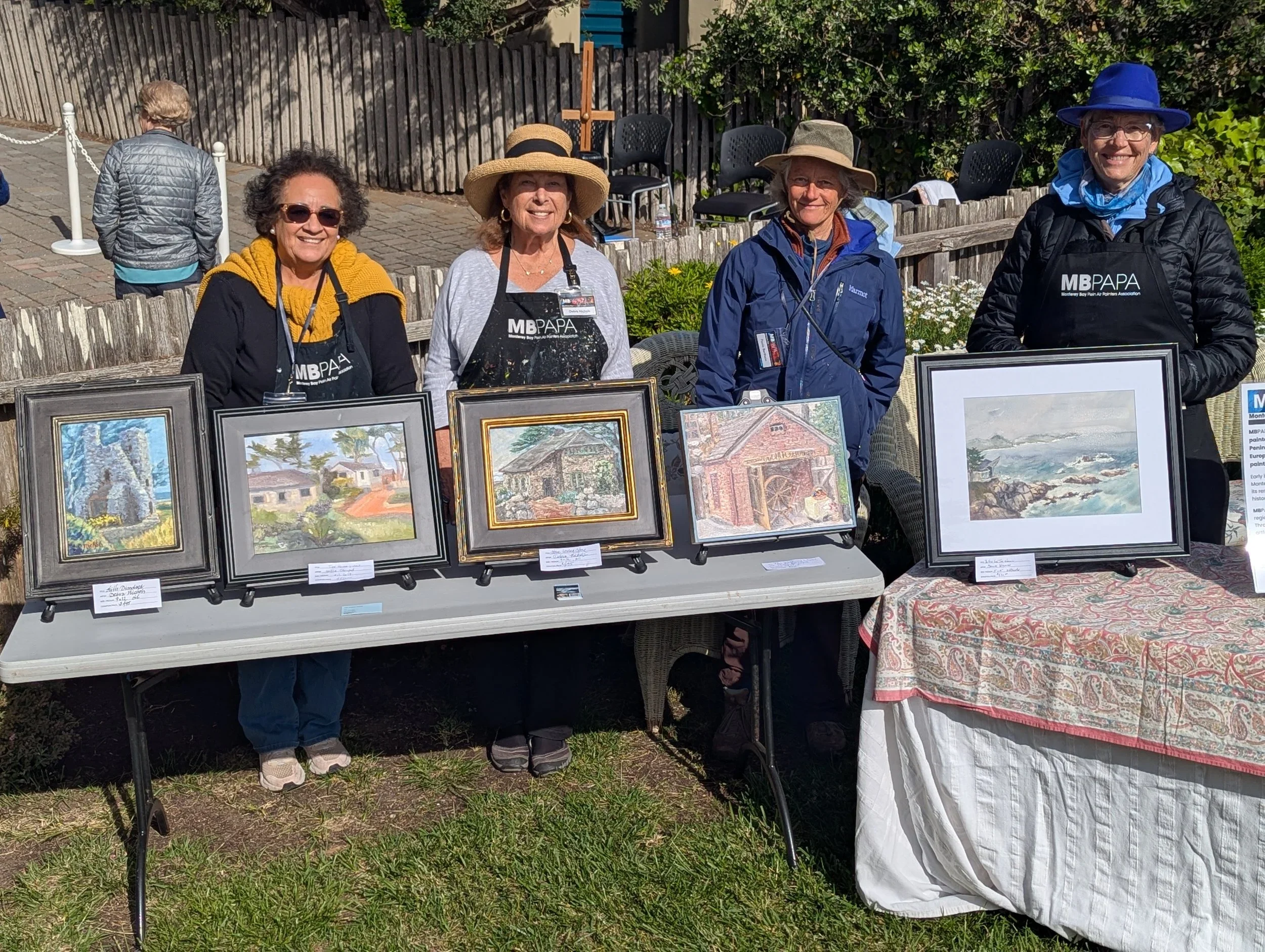 Four women standing behind display tables showcasing framed artwork. They are outdoors, some wearing hats and jackets, with a wooden fence and greenery in the background.