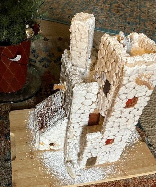 A gingerbread house under construction covered in powdered sugar, with parts of the walls and roof still missing. A Christmas tree with ornaments is visible in the background.