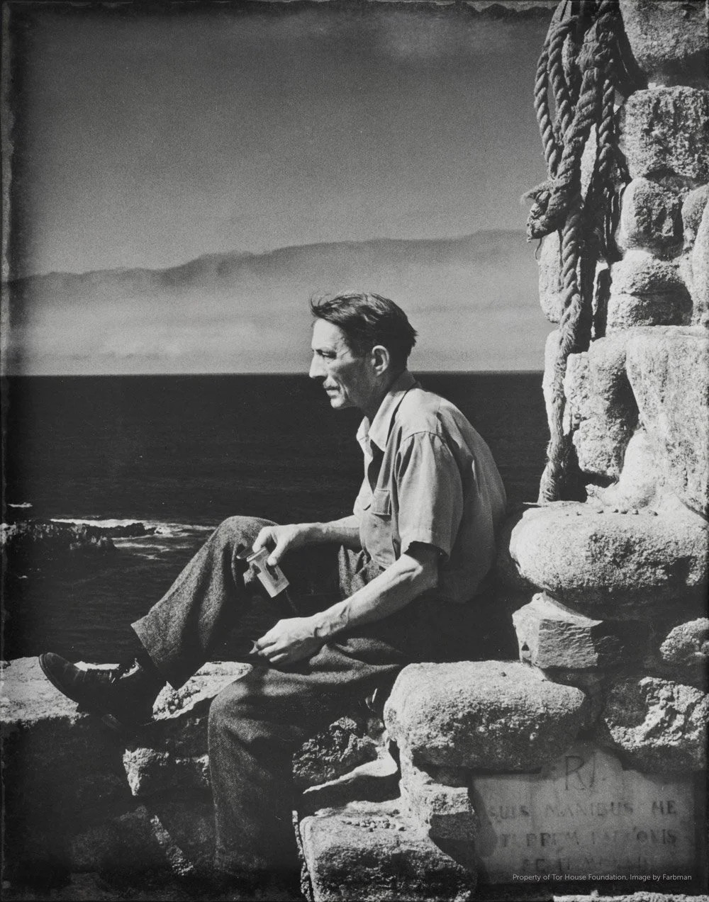 A black and white photo of a man sitting on a stone wall near the ocean with a cloudy sky in the background.