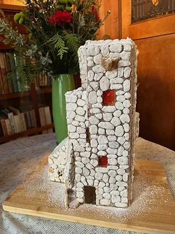 A miniature model of a stone tower with small archways and windows, placed on a wooden board. In the background, there is a green vase with red and pink flowers.