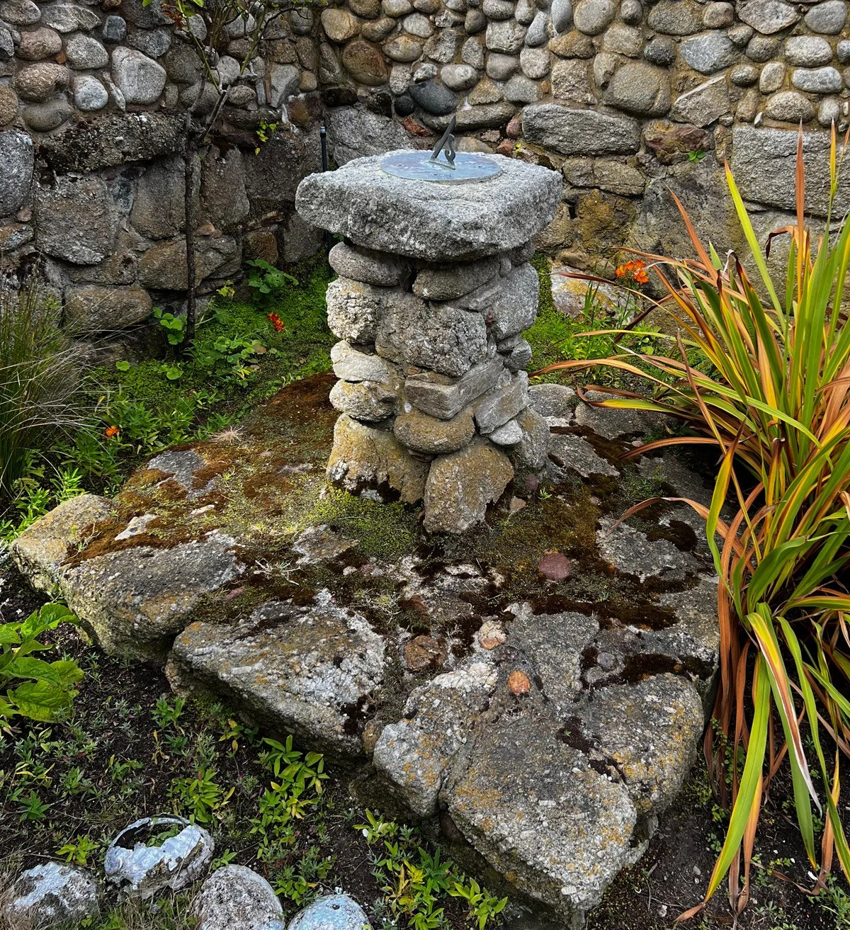 A stone sundial on a mossy ground, surrounded by plants and a stone wall in the background.
