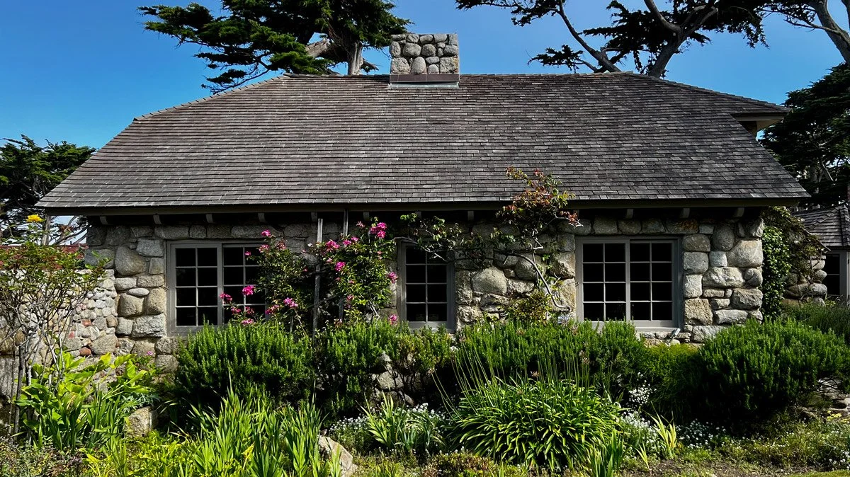 Stone cottage with a shingled roof, surrounded by lush green plants and flowering bushes, under a clear blue sky.