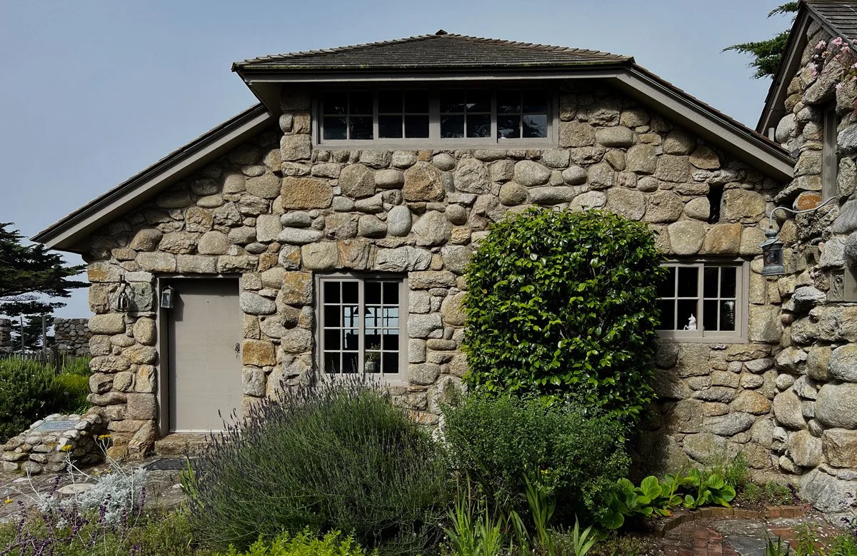 A house with a stone exterior, a gabled roof, and multiple windows, surrounded by greenery and plants.
