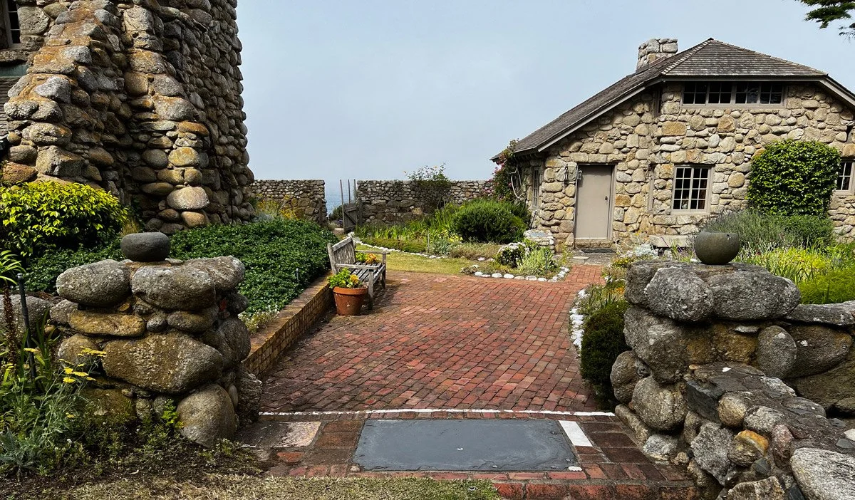 A stone house and stone wall in a garden with a brick pathway, a bench, potted plants, and landscaped greenery.