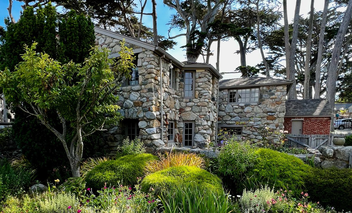 Stone house with multiple windows, surrounded by lush garden with trees and colorful plants.
