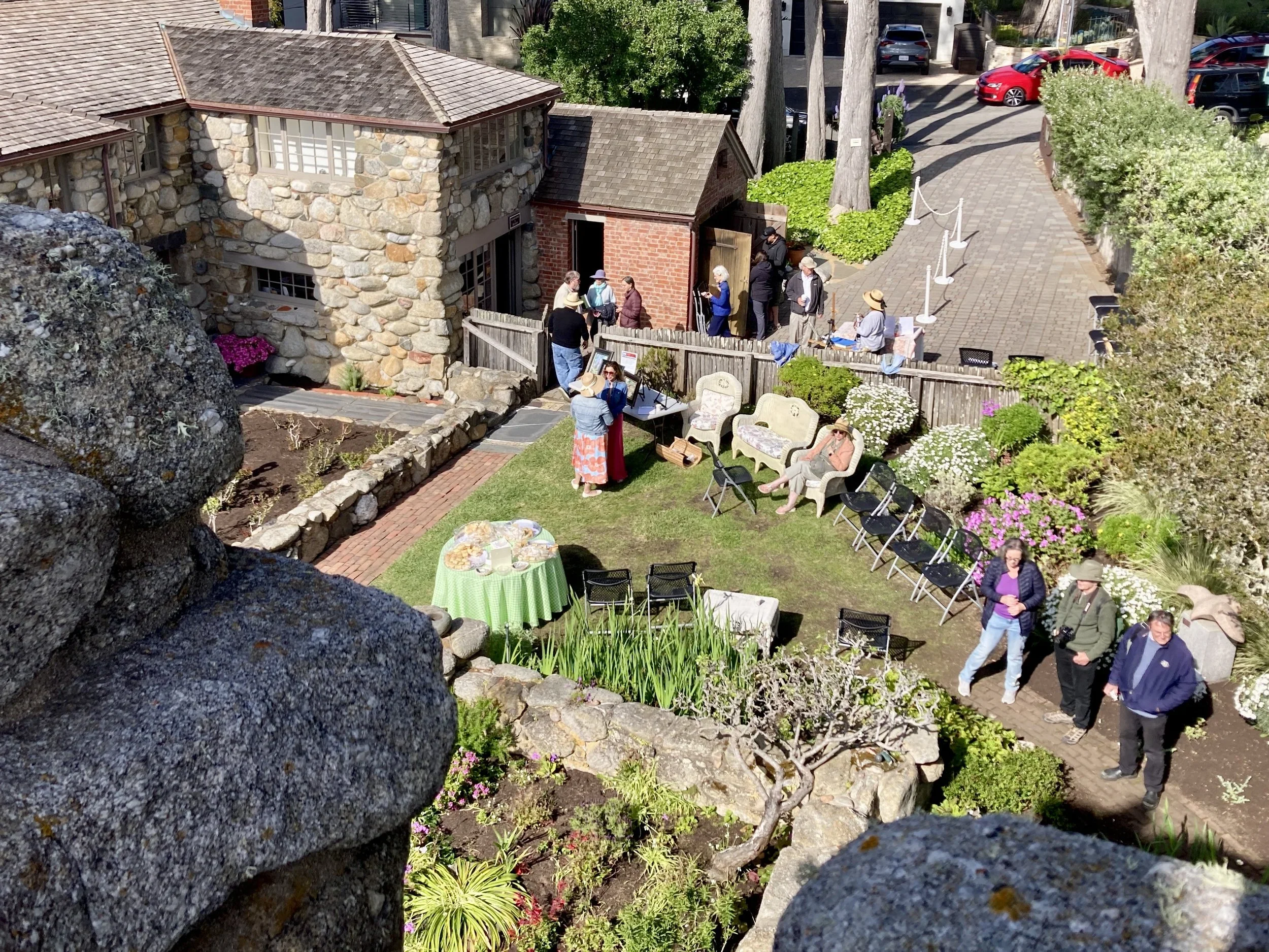 People gather in a garden courtyard with chairs and tables, some sitting and chatting, others standing and socializing. There are blooming flowers, bushes, and a stone and brick building in the background. The scene appears to be a social event or ga
