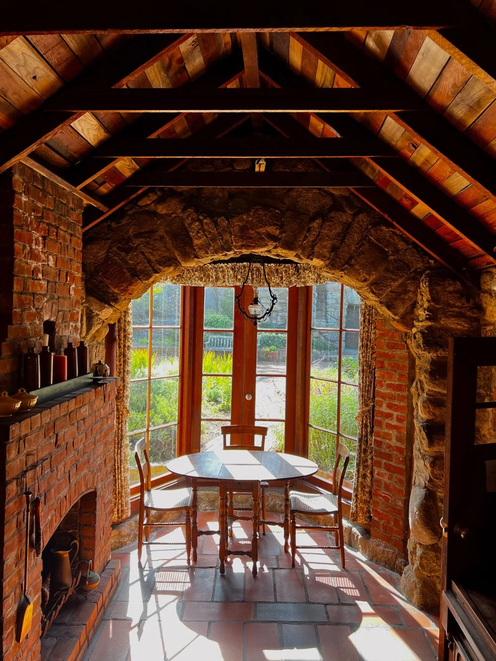 Sunlit dining area with a table and chairs, brick and stone walls, and wooden ceiling beams.