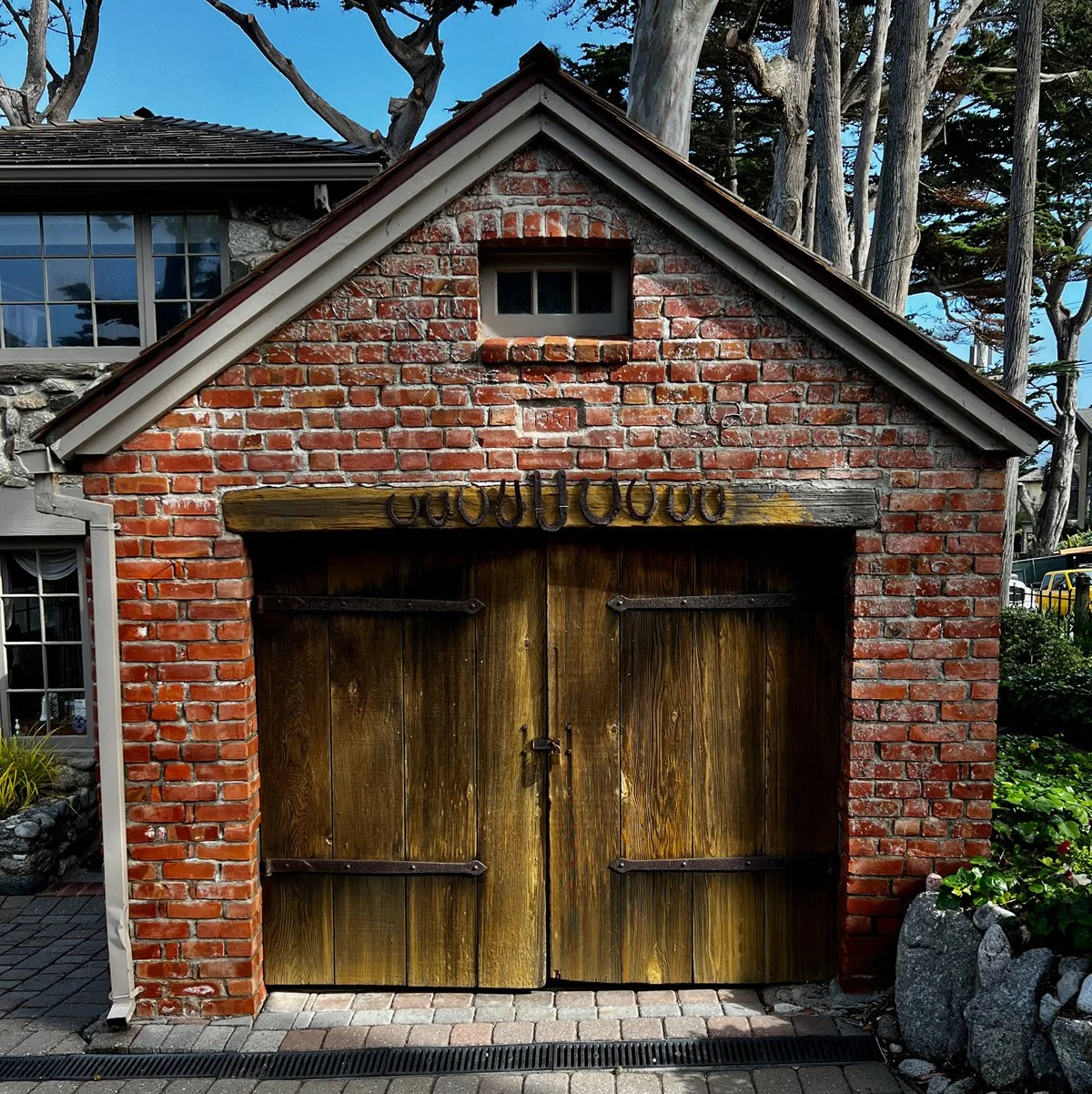 A small brick garage with wooden double doors and a wooden beam decorated with horseshoes and a small window above.