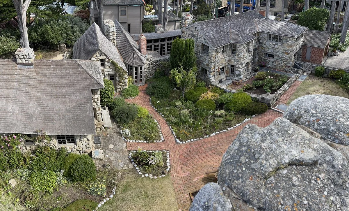 Aerial view of a quaint stone house and garden with brick pathways, surrounded by trees and other houses.