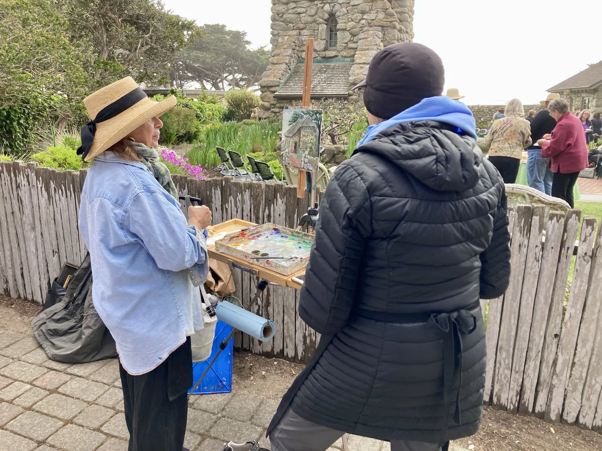 Two women stand in front of a wooden fence, one wearing a large straw hat and painting on a small easel, while the other observes. In the background, people gather outdoors near a stone building with trees and greenery.
