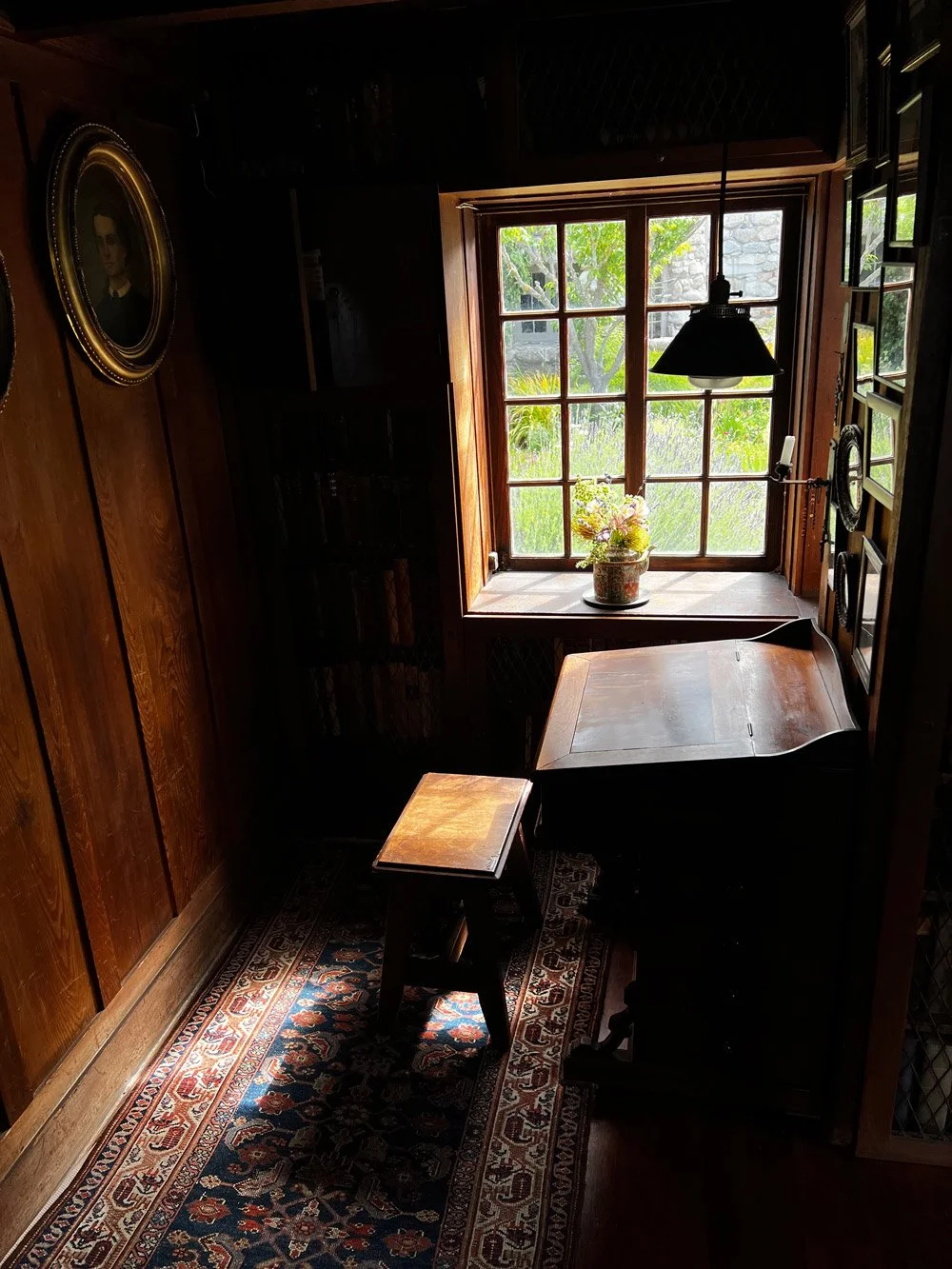 A cozy, warmly lit corner of a room featuring a wooden desk, a small wooden stool, a window with a view of green trees and grass outside, a flowerpot on the windowsill, a black hanging lamp, and a framed portrait on the wooden-paneled wall.