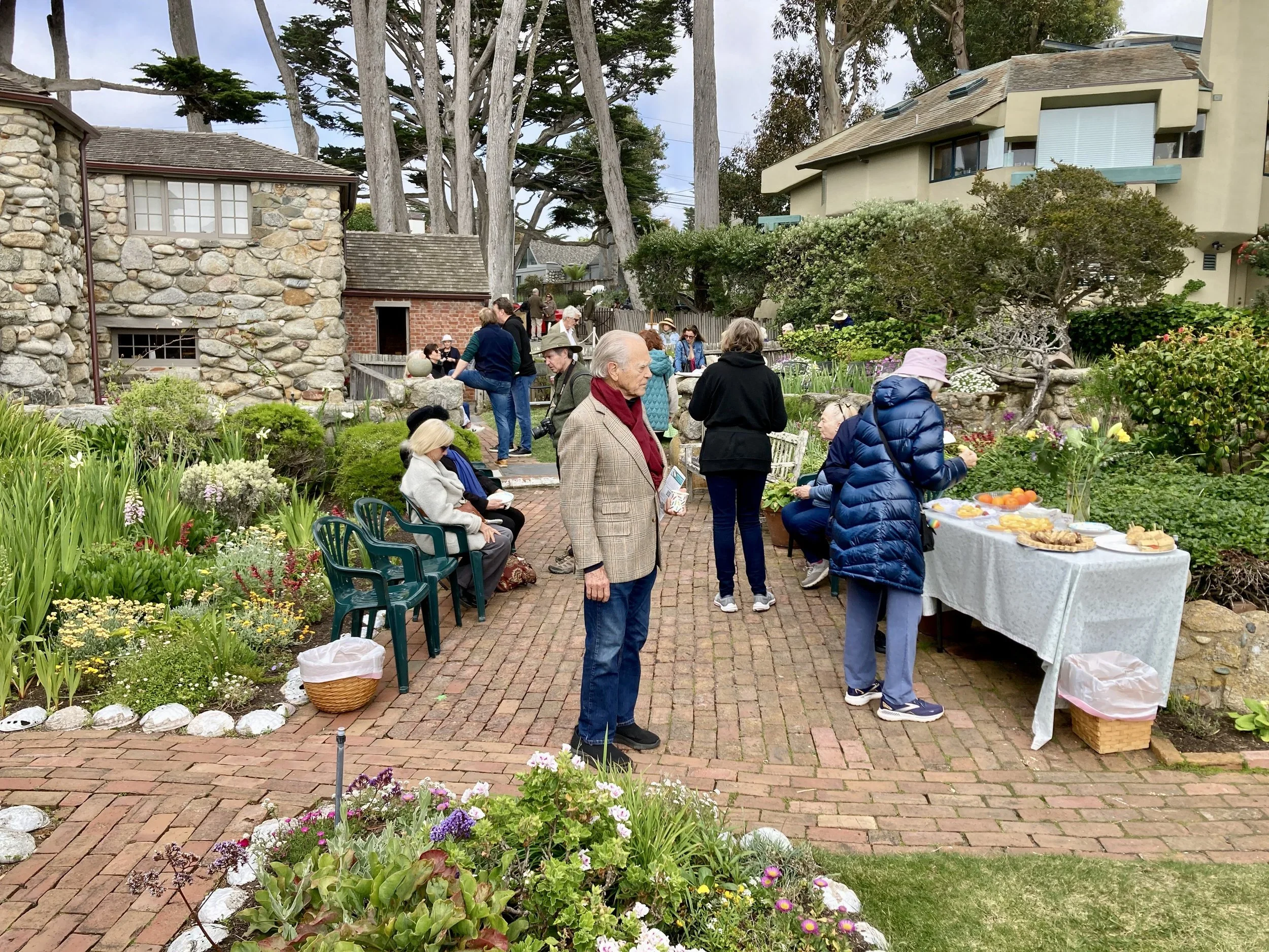 People gathering for an outdoor event in a garden with a table of food, chairs, and lush greenery in a residential neighborhood.
