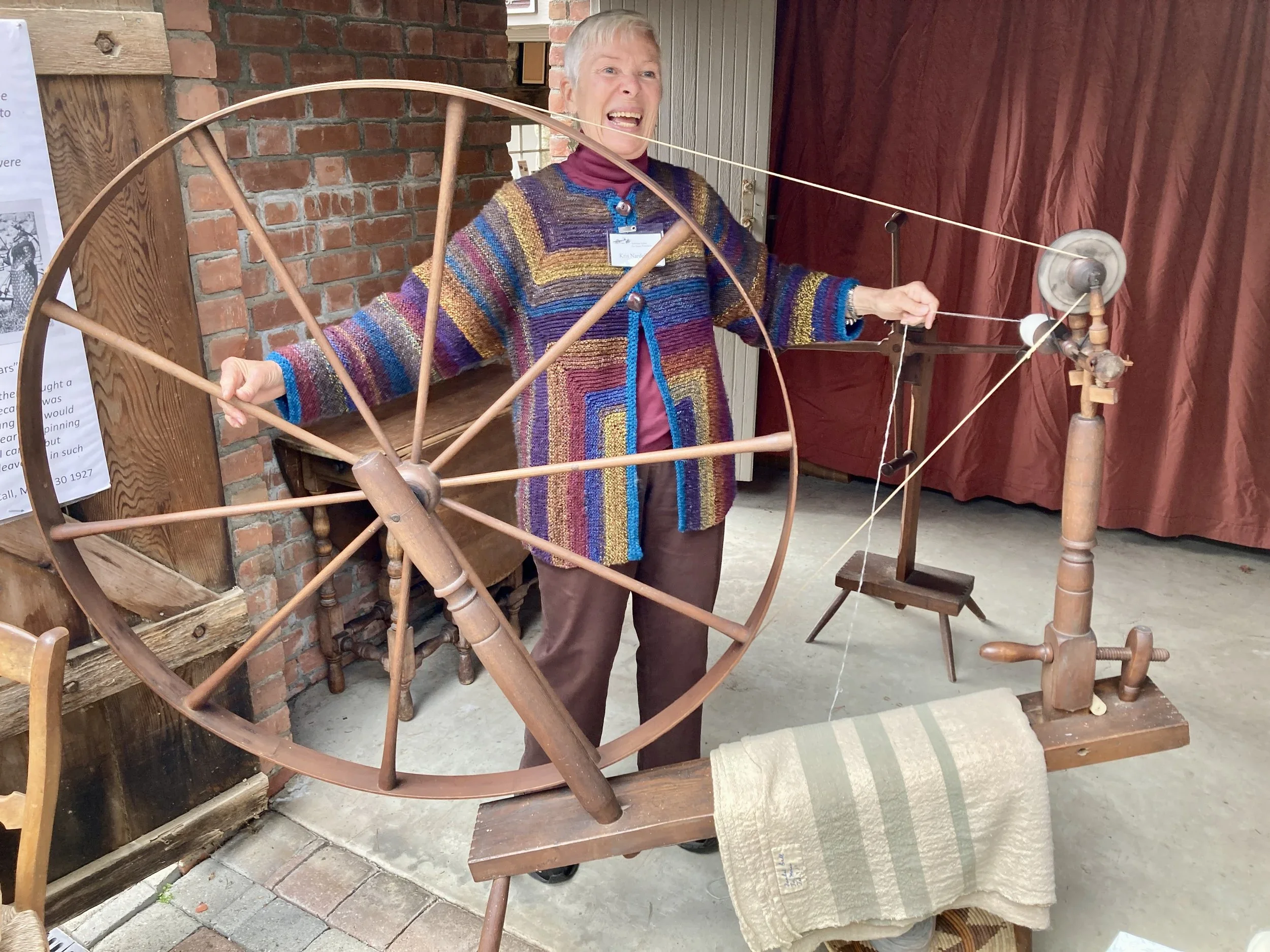 A woman operating a vintage spinning wheel with large wooden wheel and mechanical parts, standing indoors near brick wall and wooden furniture.