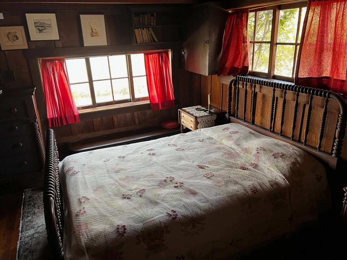 A bedroom with wooden panel walls, a bed with a floral quilt, two windows with red curtains, and a small nightstand.