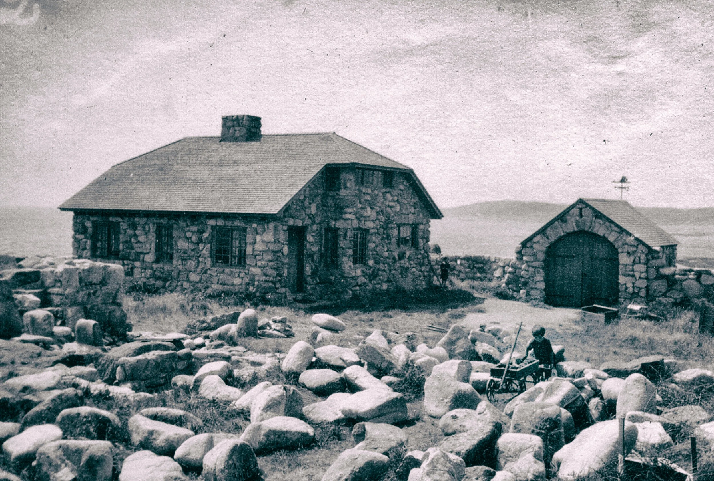 Black and white photo of a stone house and a small stone garage with a cross on top, in a rural setting with rolling hills in the background. A person is pushing a wheelbarrow among scattered rocks.