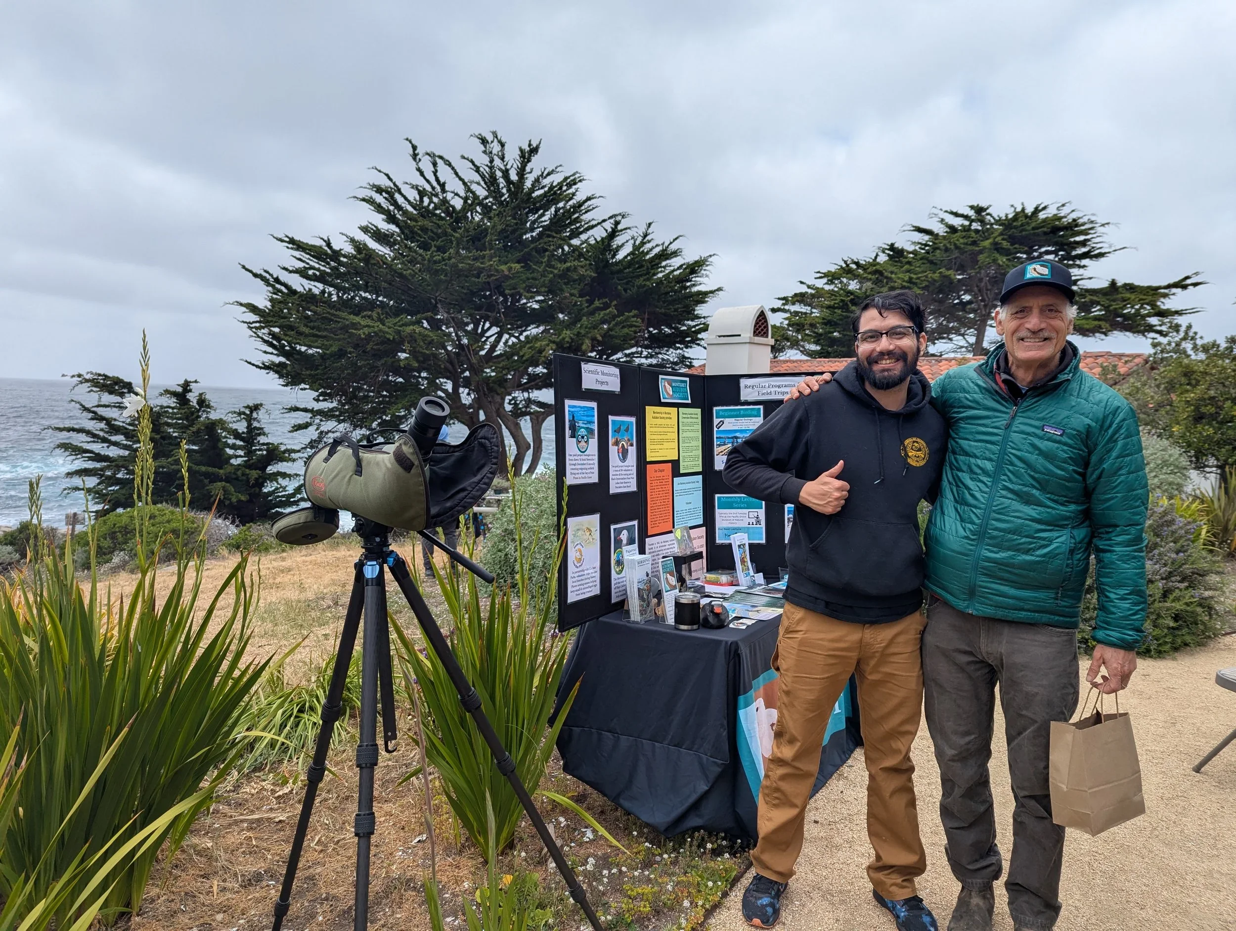 Two men standing outdoors near an informational display board on a cloudy day. One man wears a black hoodie and the other a teal jacket, and they are smiling and posing for the camera. A telescope is set up on a tripod nearby, and the background feat