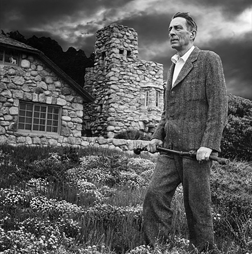 A man with a serious expression holding a gardening tool in front of a stone house with a chimney and mountains in the background, under a stormy sky.