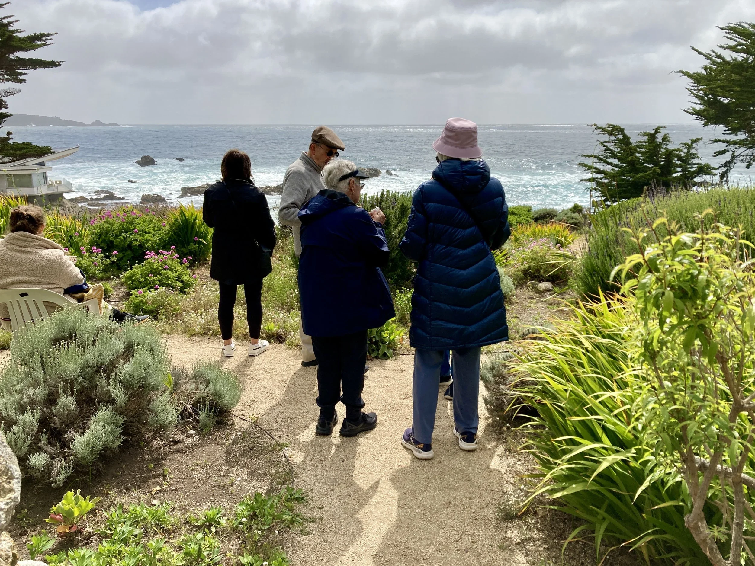 Group of six people on a coastal garden path looking towards the ocean on cloudy day.