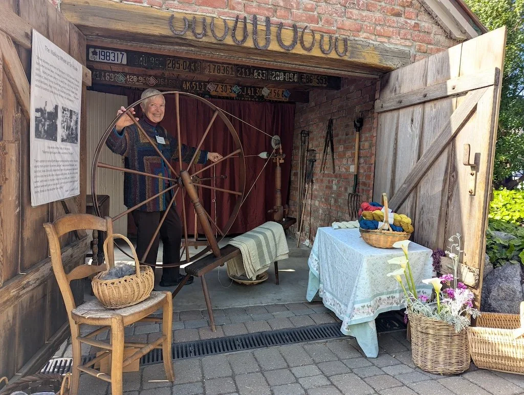 A woman standing inside a rustic outdoor workshop with a spinning wheel, surrounded by baskets of yarn, tools, and floral arrangements.