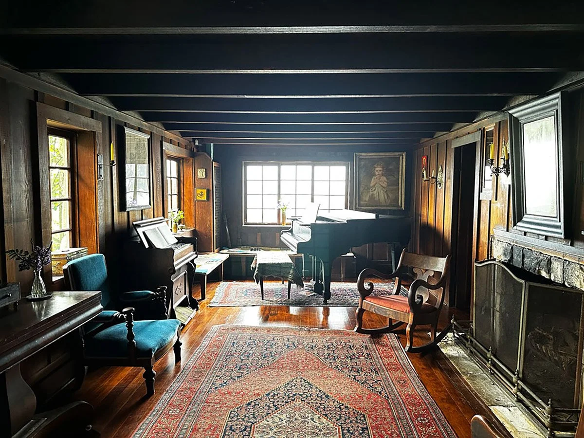 A cozy room with wooden panel walls, large windows, a fireplace, vintage furniture, a piano, and a colorful Oriental rug.