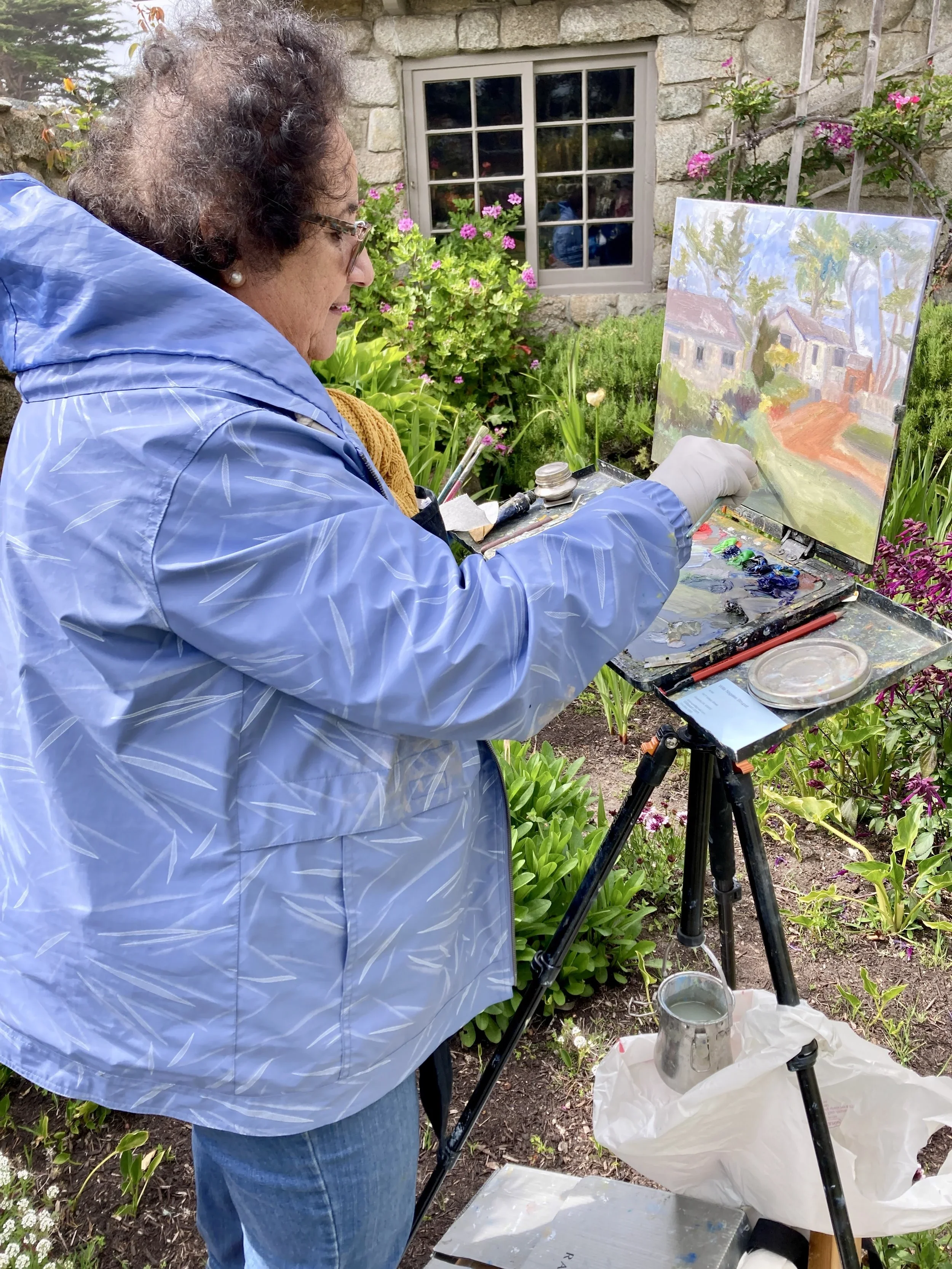 An elderly woman painting outdoors in a garden with various pink and purple flowers, a stone house wall, and a window in the background.